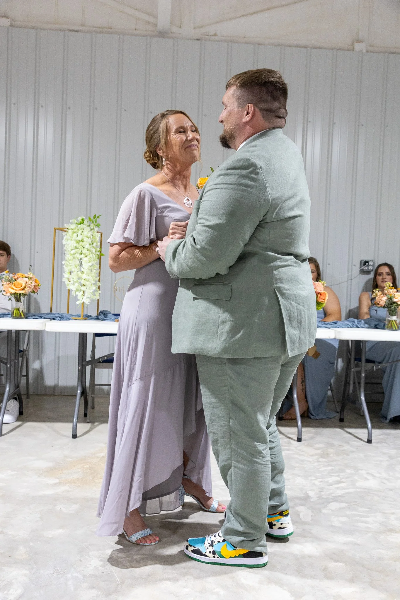 A woman and a man dancing together at a celebration, with women sitting at a table with flower arrangements in the background.