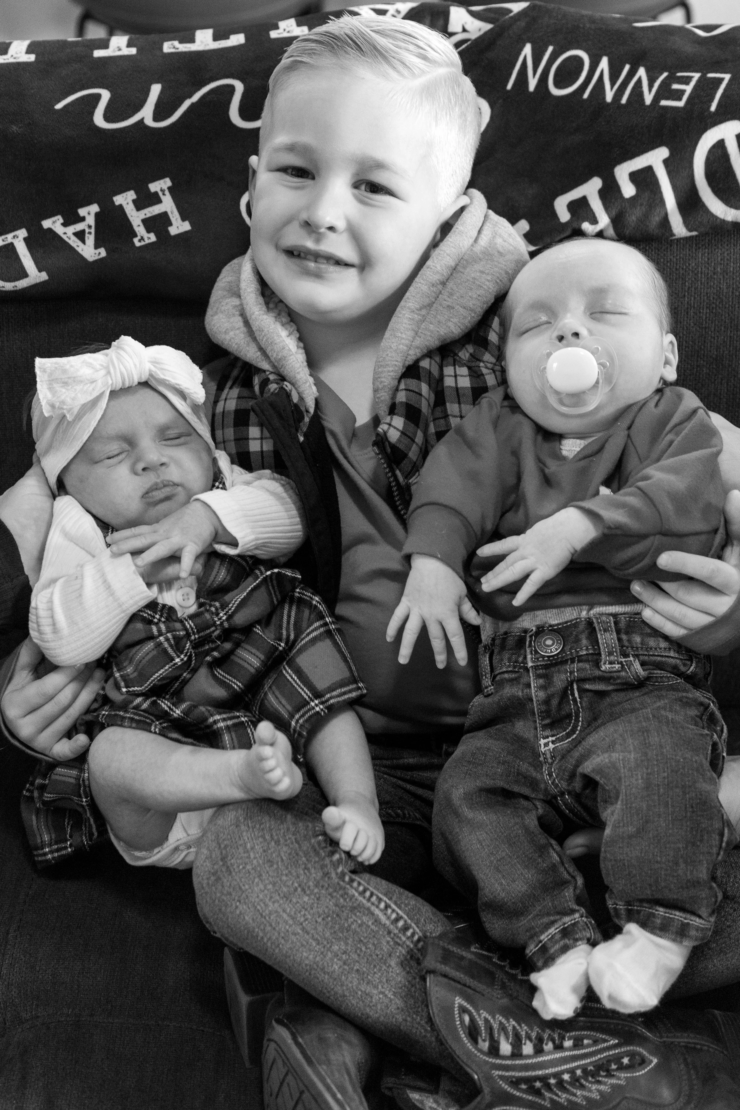 A young boy sitting on a couch holding two sleeping babies, one in each arm, with a blanket and pillows behind them.