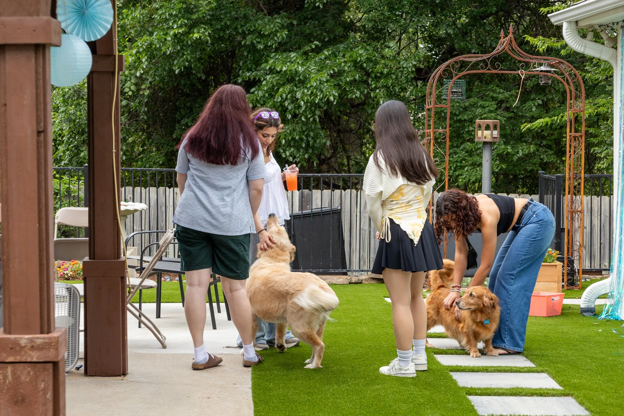 Four women and two dogs in a backyard, with one woman petting a dog, and the others observing, surrounded by green trees and outdoor furniture.