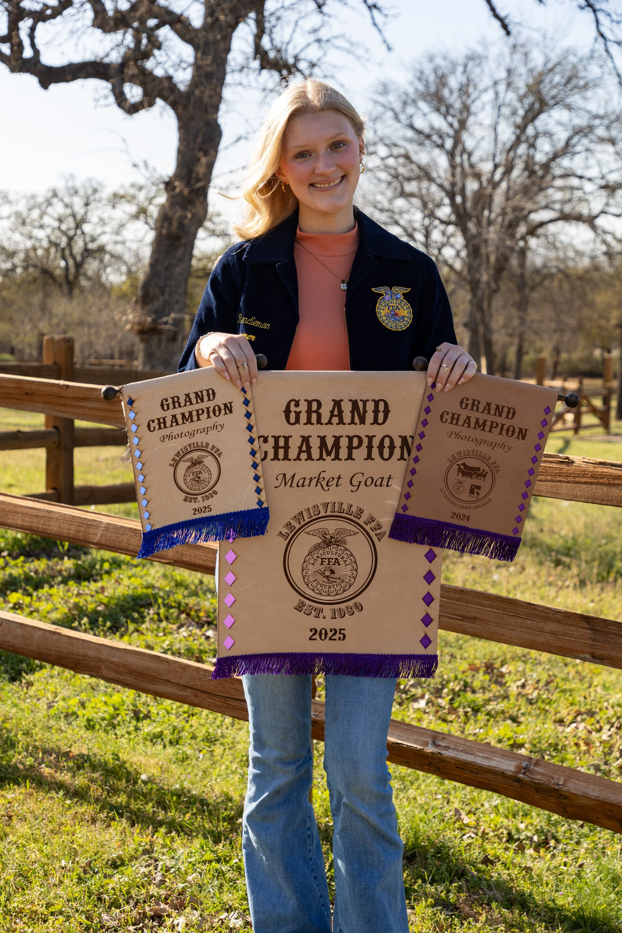 A young woman standing outdoors on a farm, holding three awards for Grand Champion Market Goat, with trees and a wooden fence in the background.