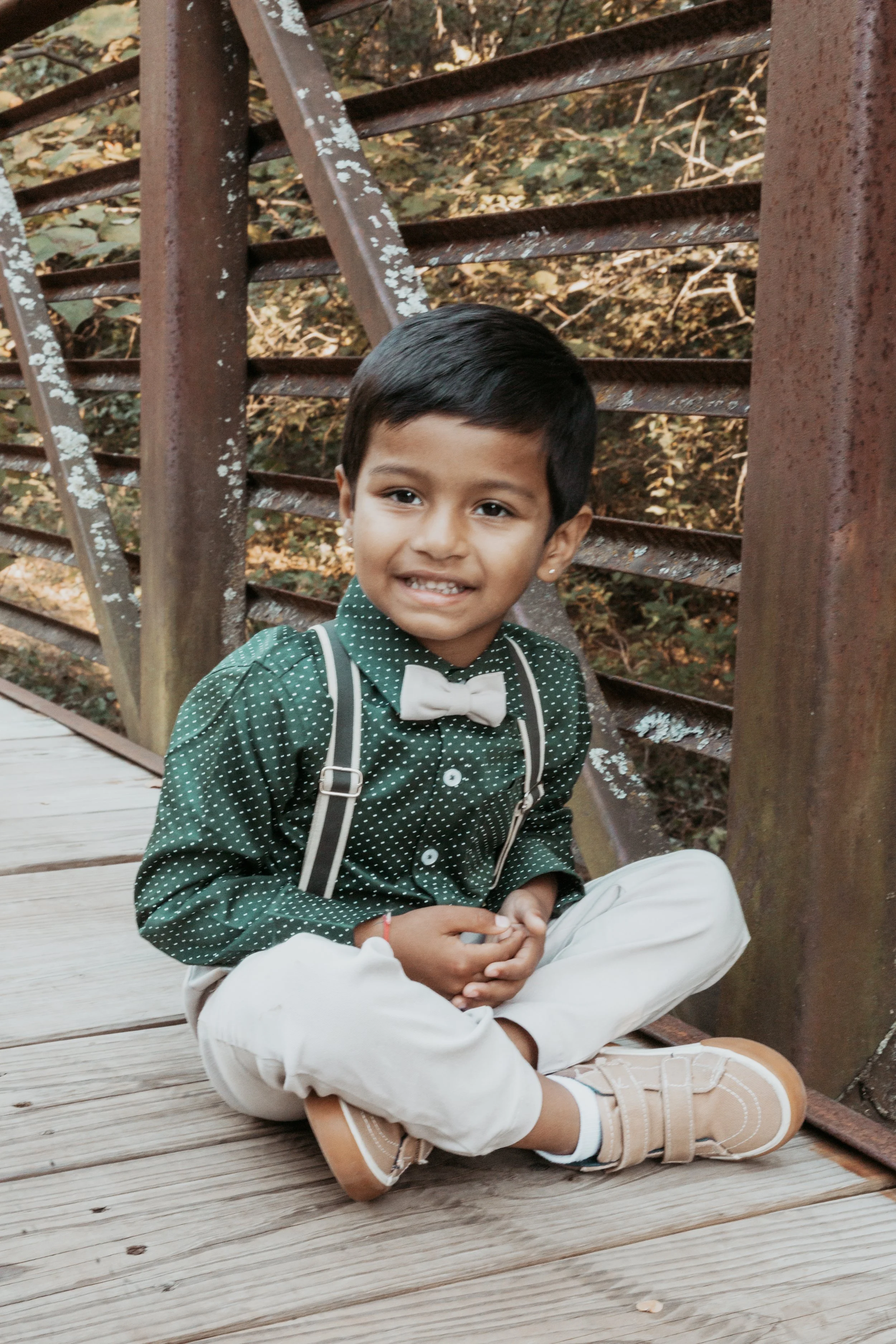 A young boy sitting cross-legged on a wooden bridge, wearing a dark green shirt with white polka dots, a white bow tie, beige pants, and tan shoes, smiling at the camera.
