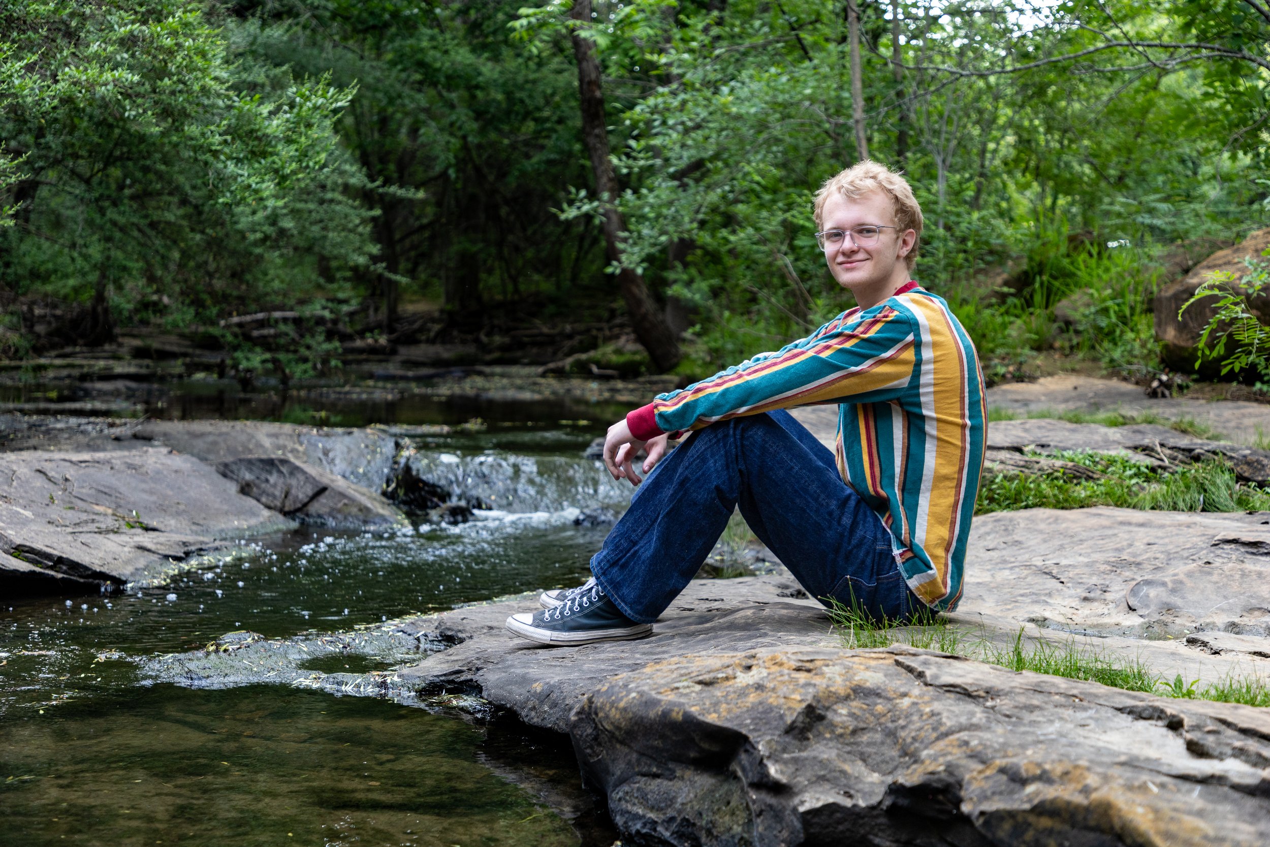 A young person with blonde hair, glasses, wearing a colorful striped shirt, jeans, and sneakers, sitting on a large rock by a small creek in a forested area.