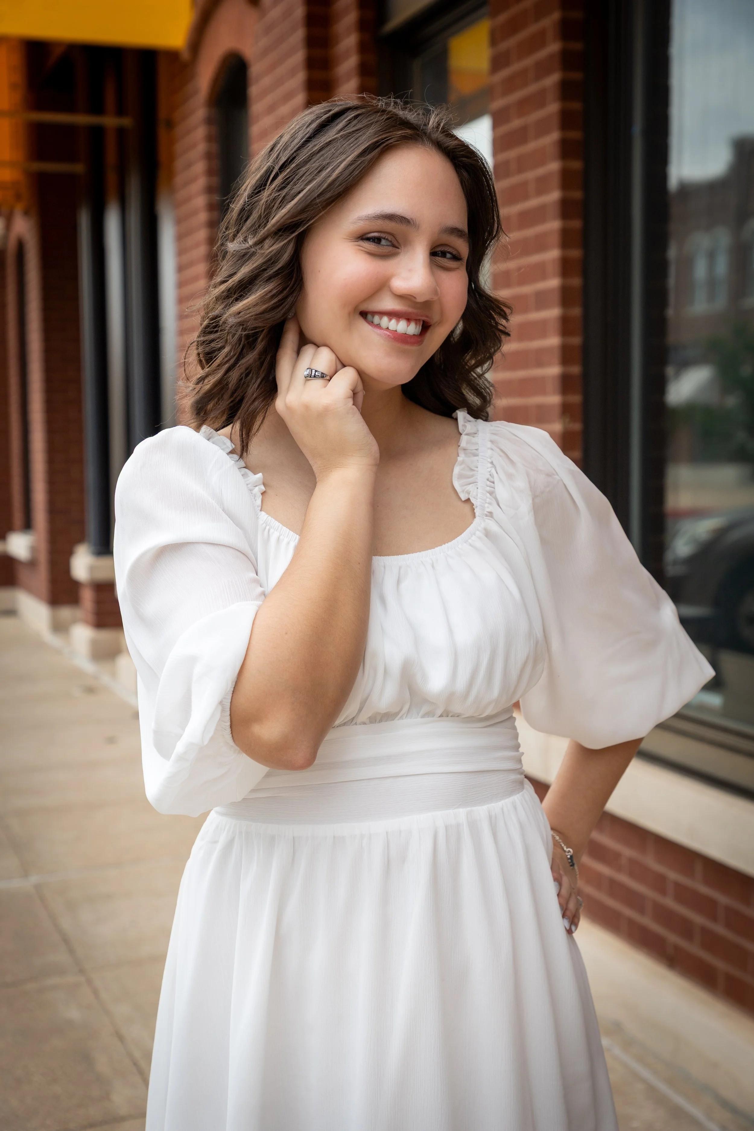 Young woman with brown hair styled in loose waves, smiling, wearing a white dress with puffed sleeves, standing outdoors against a brick building with large windows.