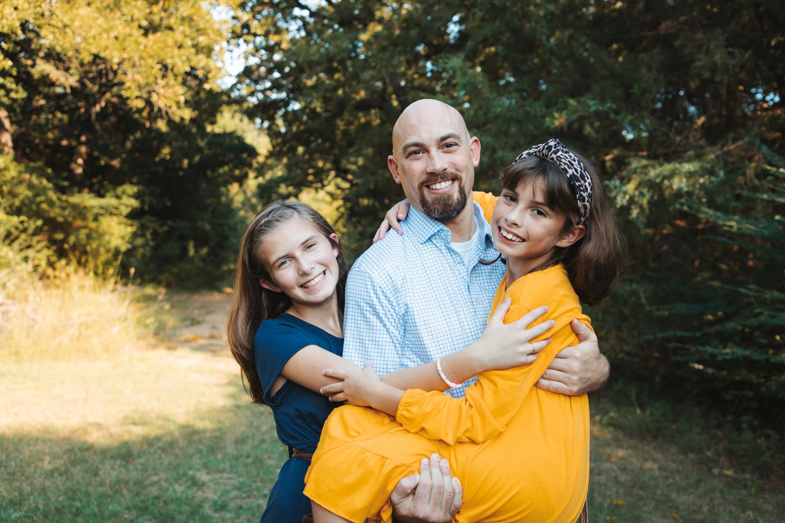 A man holding two smiling young girls outdoors in a park with trees in the background.