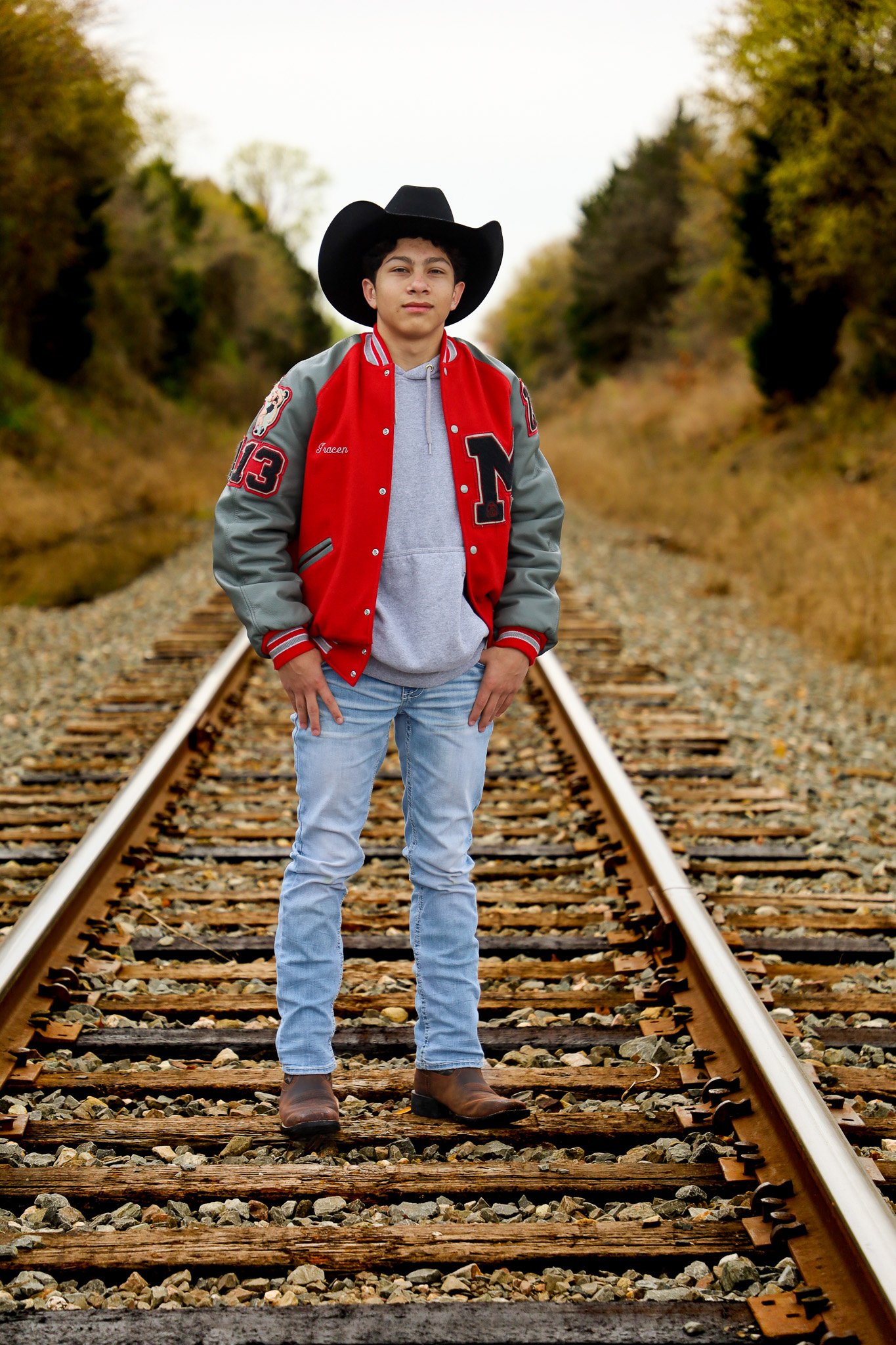 Young man standing on train tracks wearing a black cowboy hat, red and gray varsity jacket, light blue jeans, and brown boots, with autumn foliage in the background.