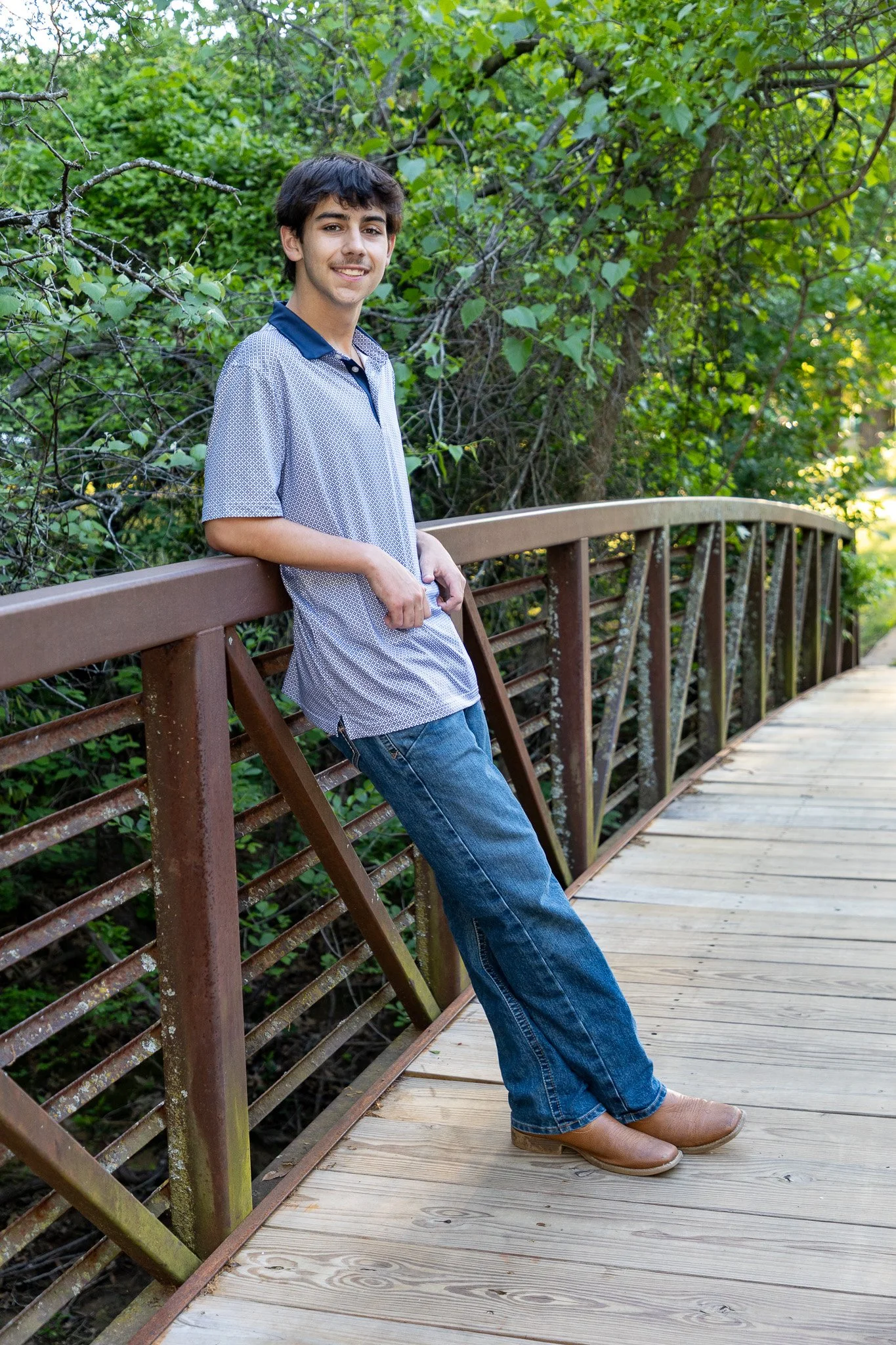 A young man in a short-sleeved, patterned shirt and jeans leaning against a brown wooden railing on a bridge, surrounded by green foliage.