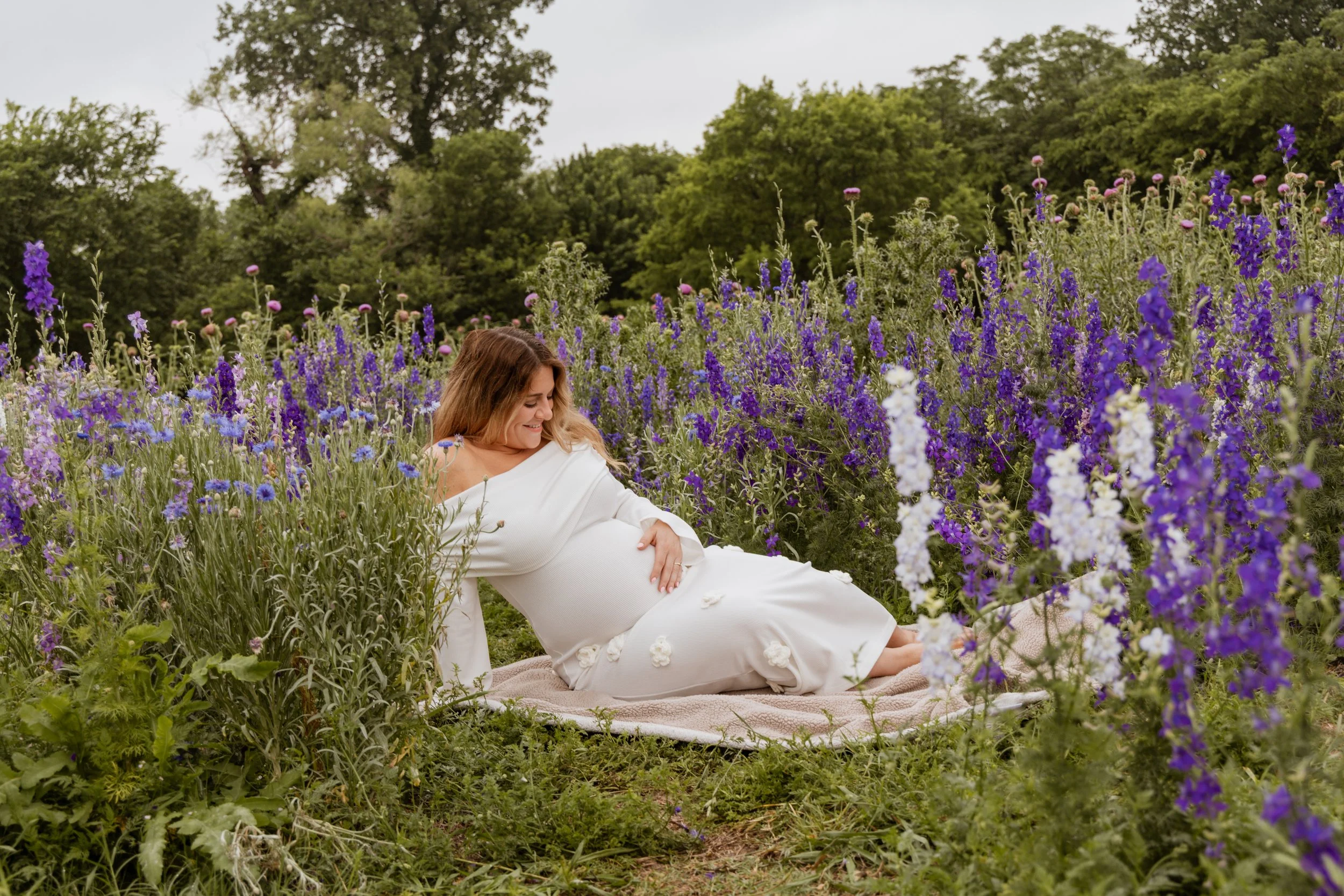 Pregnant woman lying on a blanket in a flower garden, surrounded by purple and white flowers, with trees in the background.