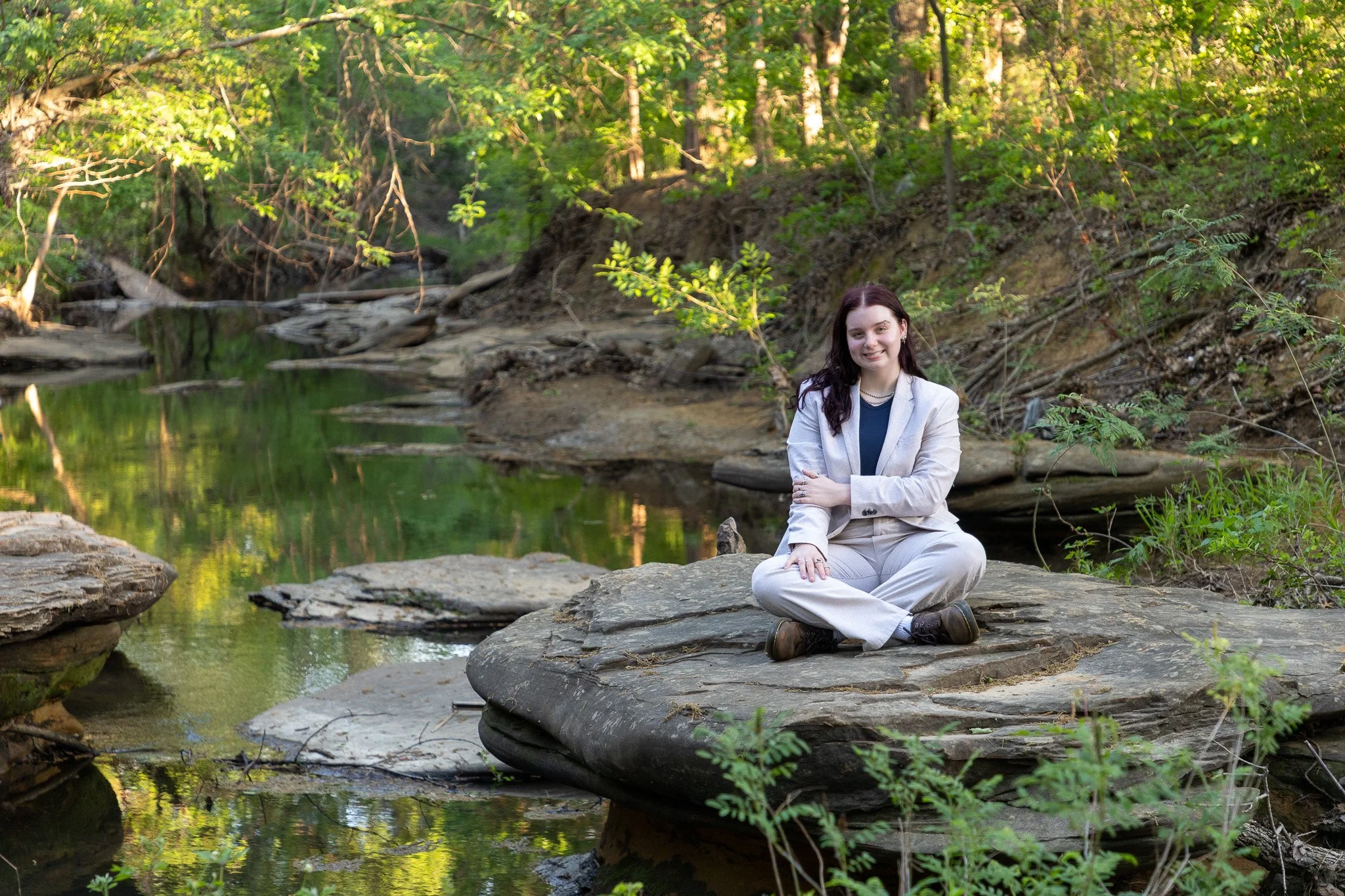 A woman in a light-colored suit sitting cross-legged on a flat rock near a stream in a lush green forest, smiling at the camera.