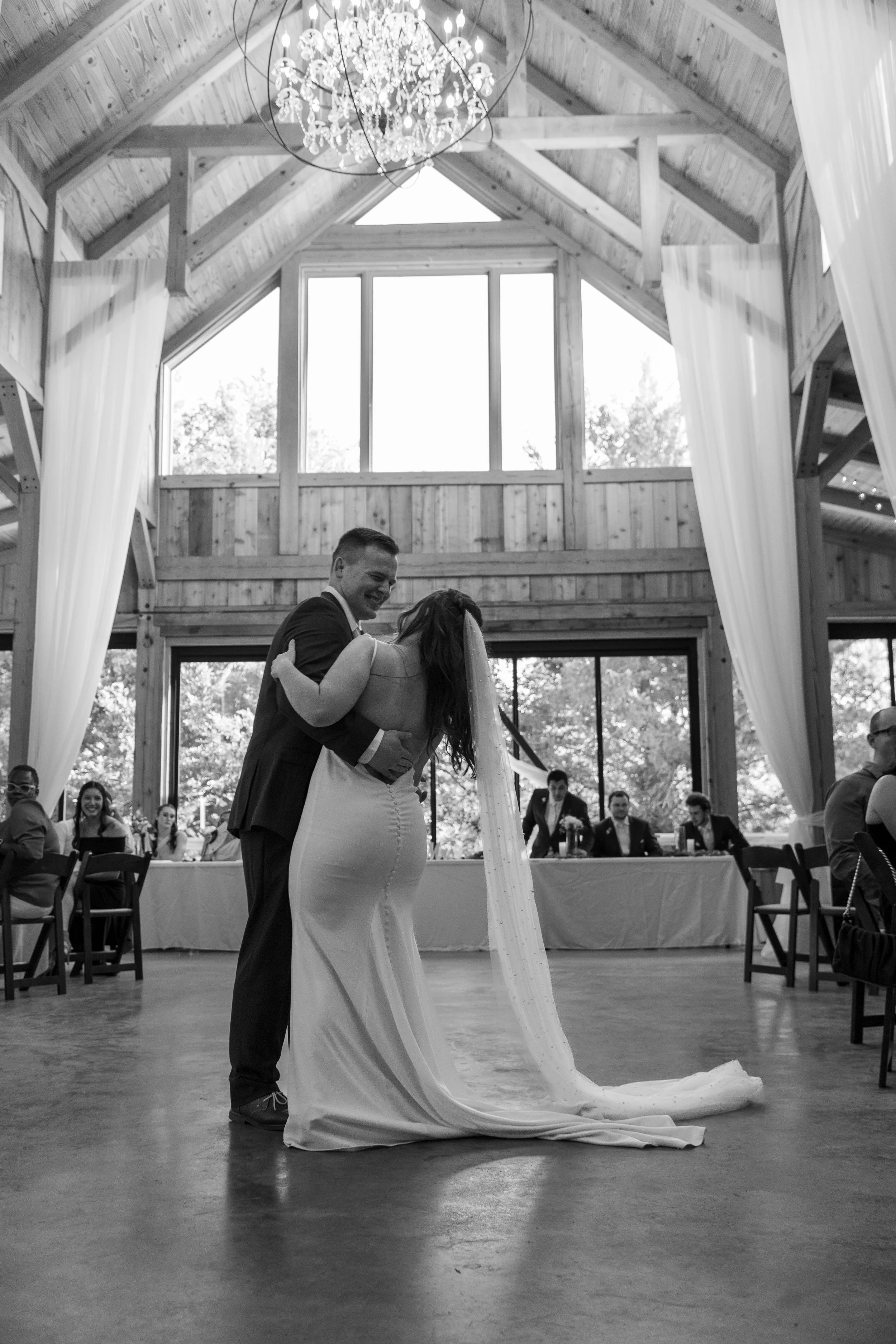 Black and white photo of a bride and groom sharing a first dance at their wedding reception inside a wooden barn with high ceiling, chandeliers, and large windows.