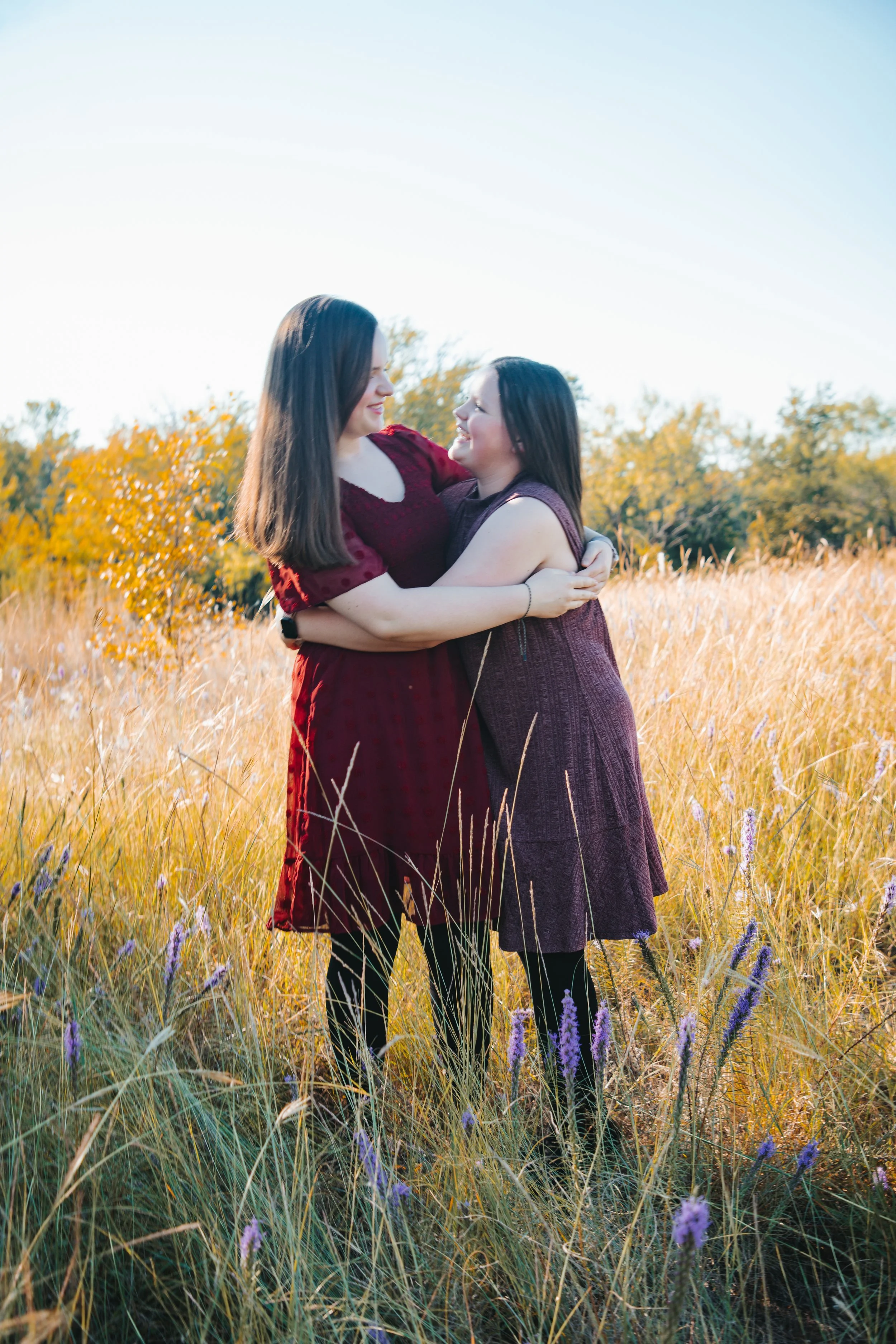Two women in red dresses hugging and smiling in a field of tall grass and wildflowers during sunset.