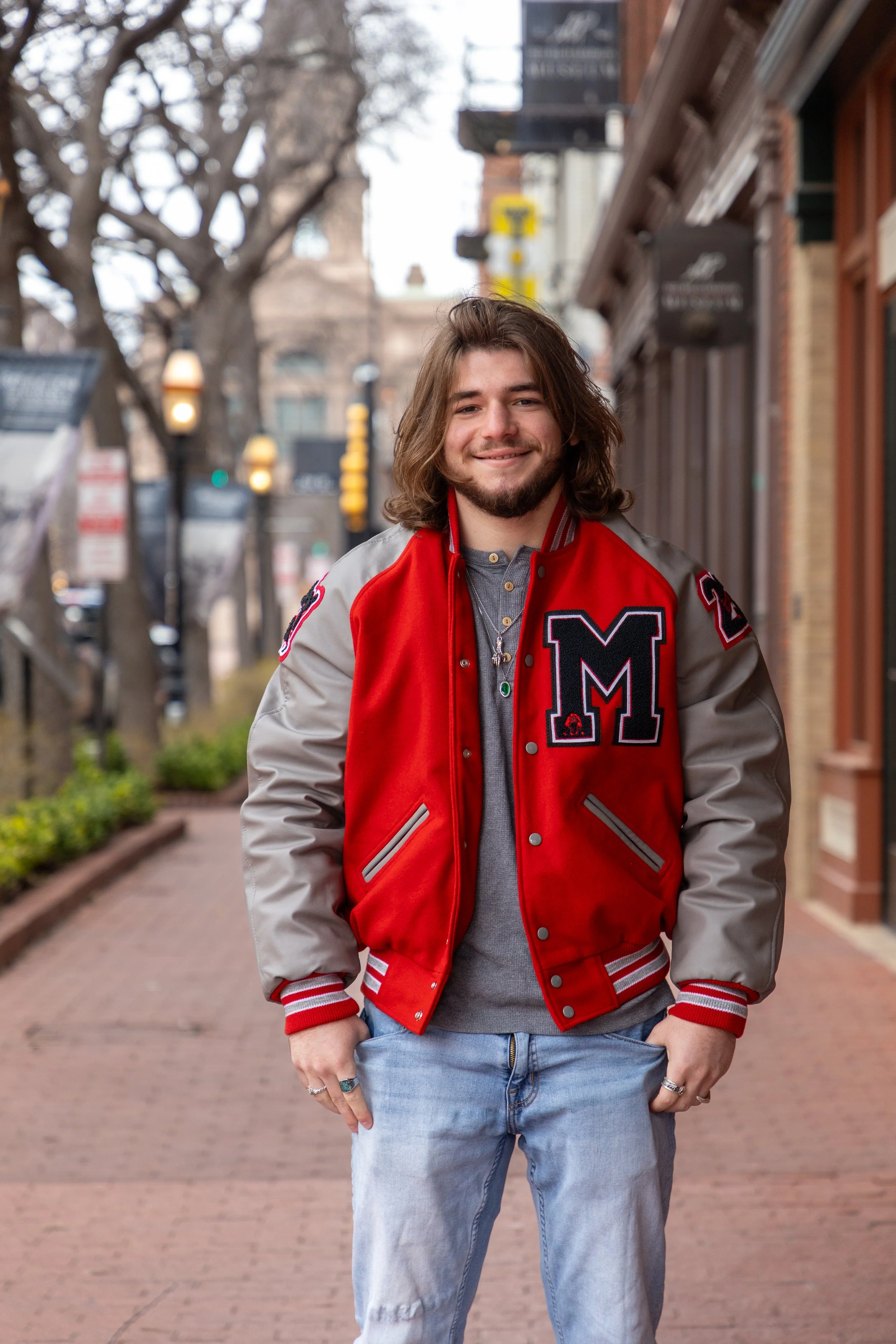 A young man with long brown hair and a beard standing on a city sidewalk, smiling, wearing a red and gray varsity jacket with an 'M' patch, gray shirt, and light blue jeans.