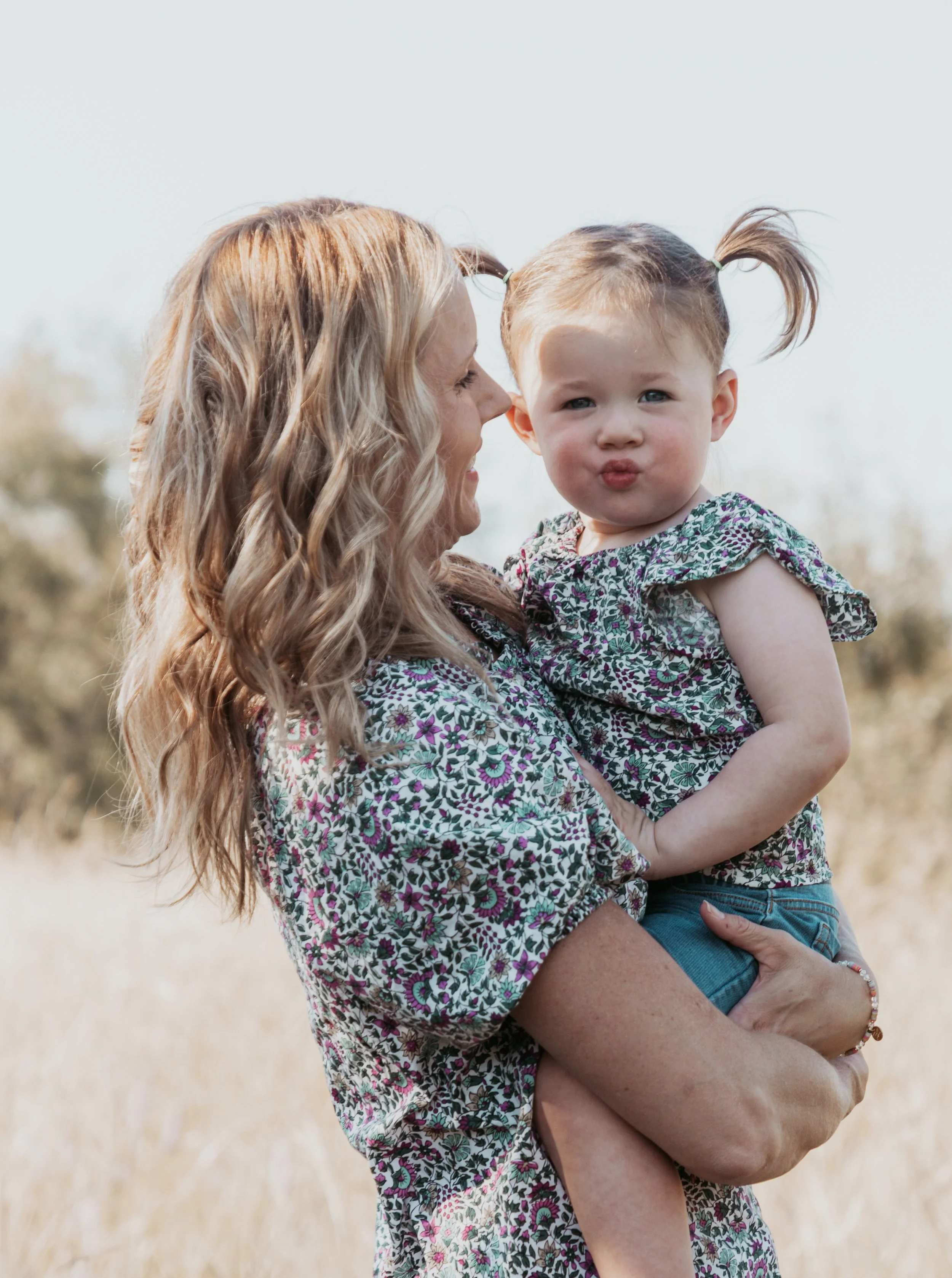 A woman with long, wavy blonde hair holding a young girl with pigtails outside in a field, the girl making a kissy face.