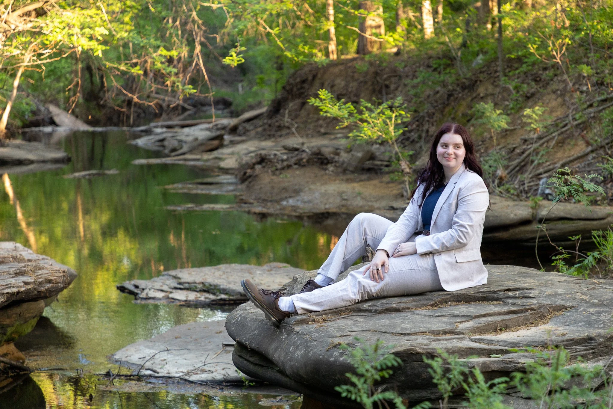 A woman with dark hair in a beige suit, blue shirt, and brown shoes, sitting on a large flat rock by a small creek surrounded by green trees and foliage.