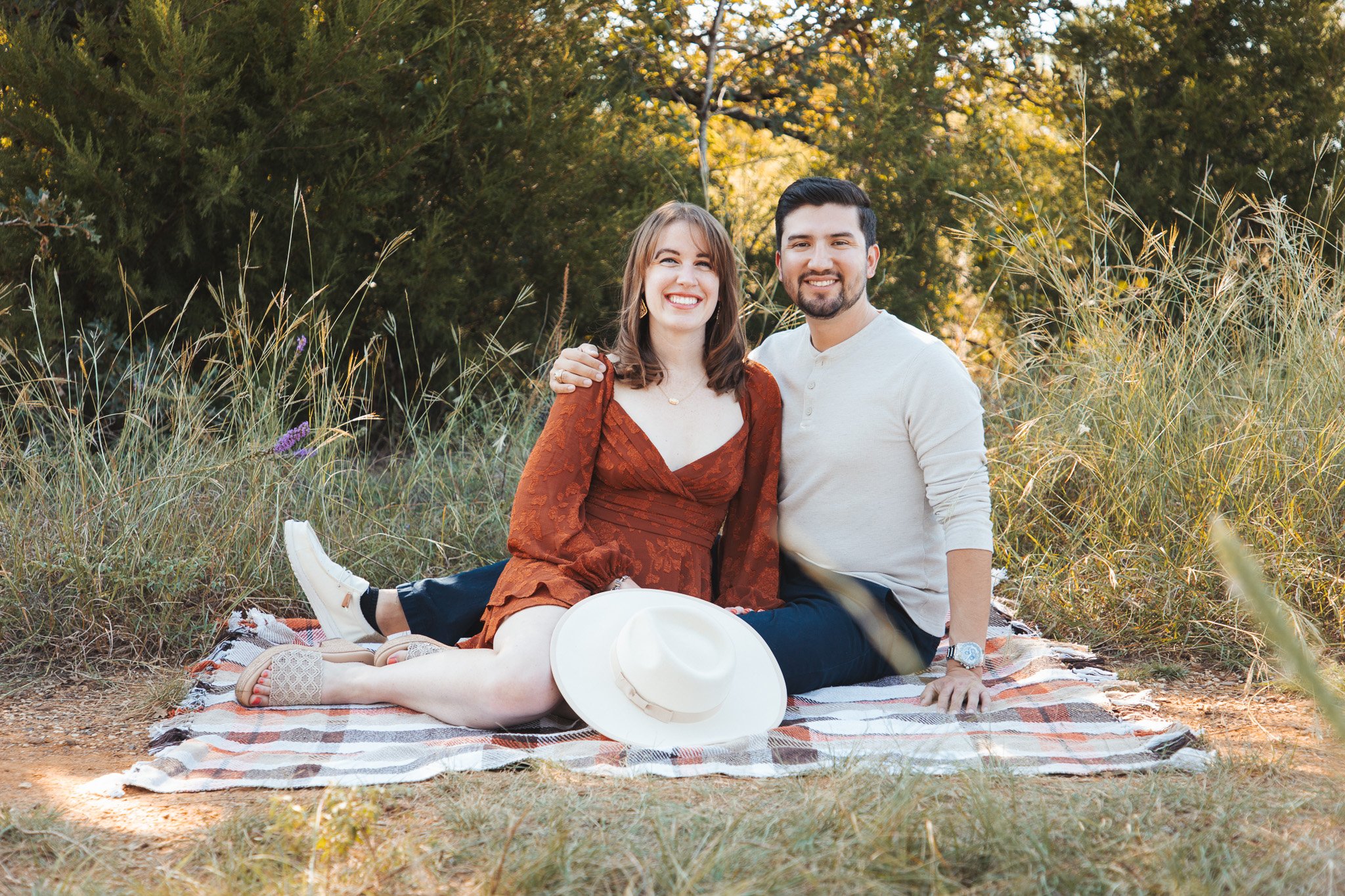 A couple sitting on a blanket outdoors, smiling with a white hat placed in front of them, surrounded by grasses and trees in the background during sunset.