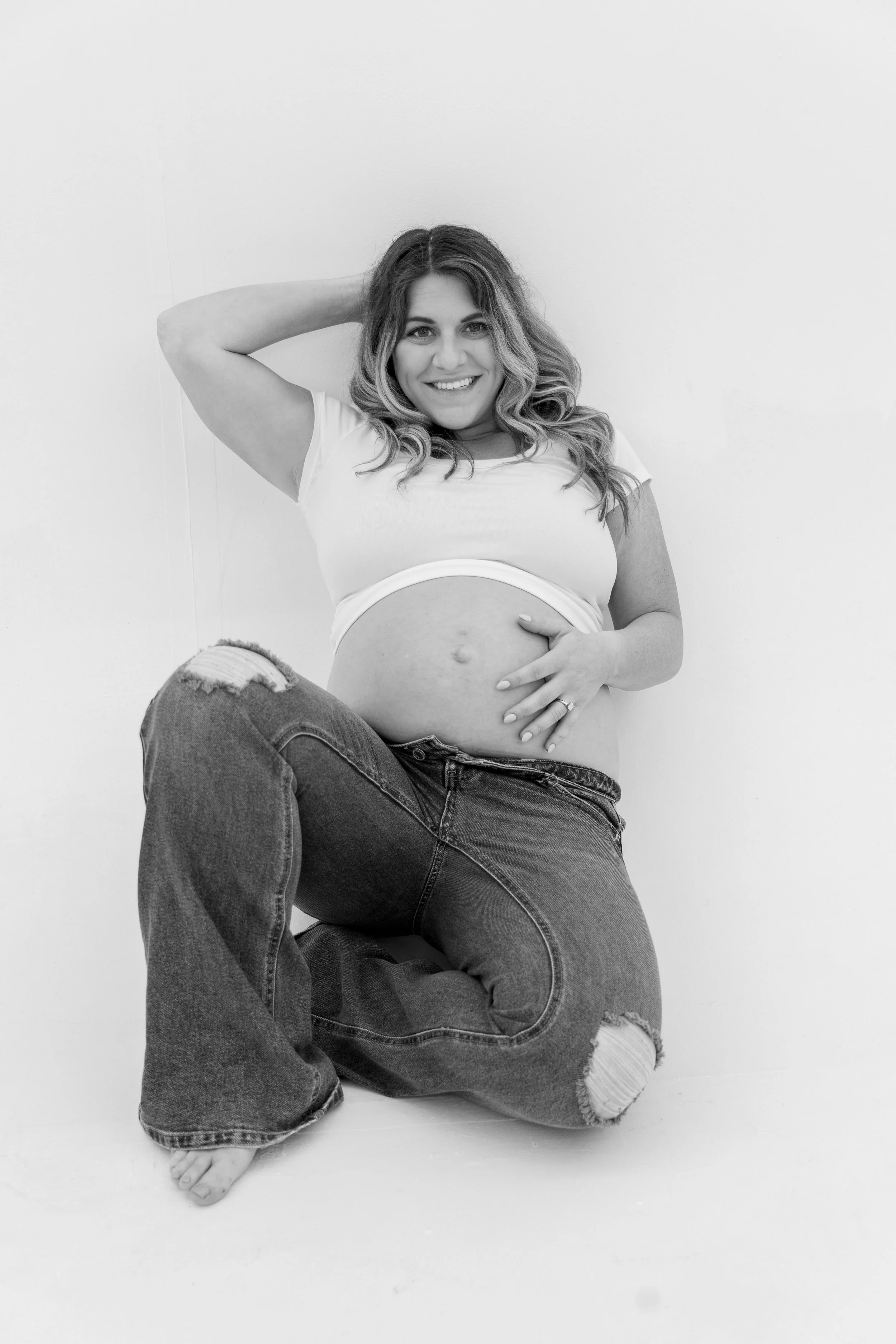 Black and white photo of a smiling woman with wavy hair, sitting on the floor against a plain wall. She is wearing a cropped t-shirt and ripped jeans, with one hand on her pregnant belly and the other resting behind her head.
