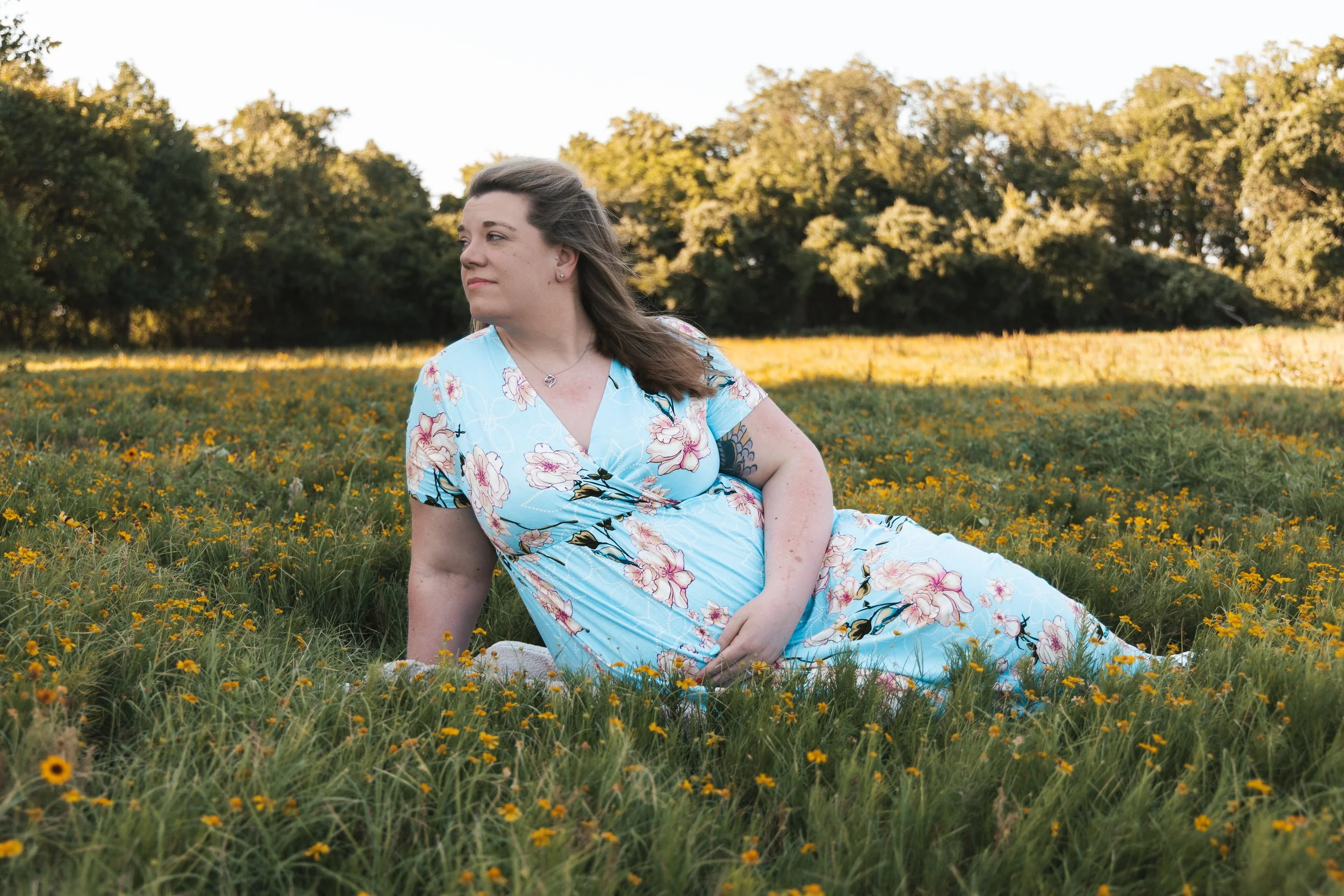 Woman in a floral dress sitting in a field of yellow flowers with trees in the background during sunset.