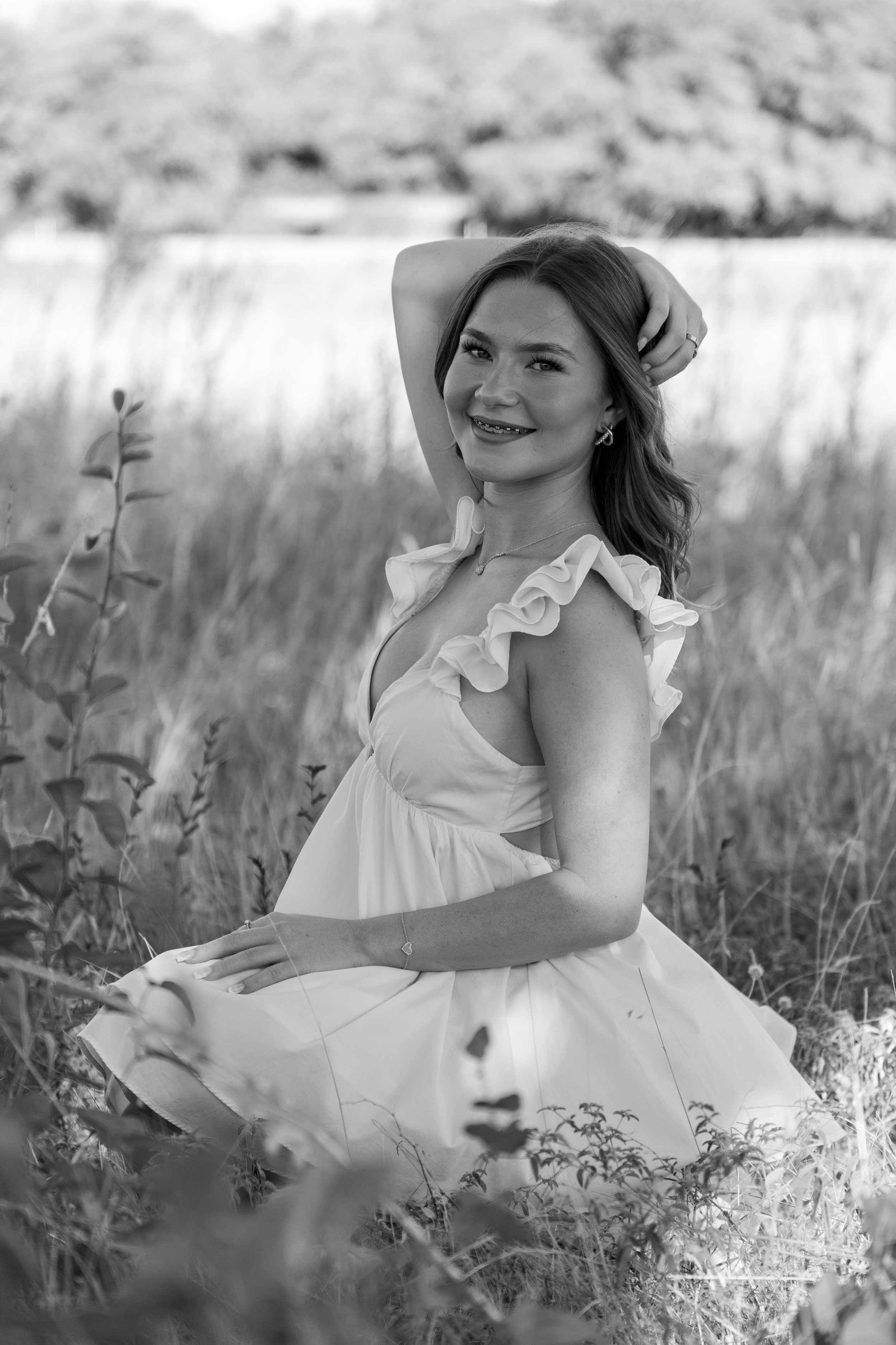 A young woman with long hair smiling and sitting in a grassy field, wearing a dress with ruffled straps. Black and white photo.