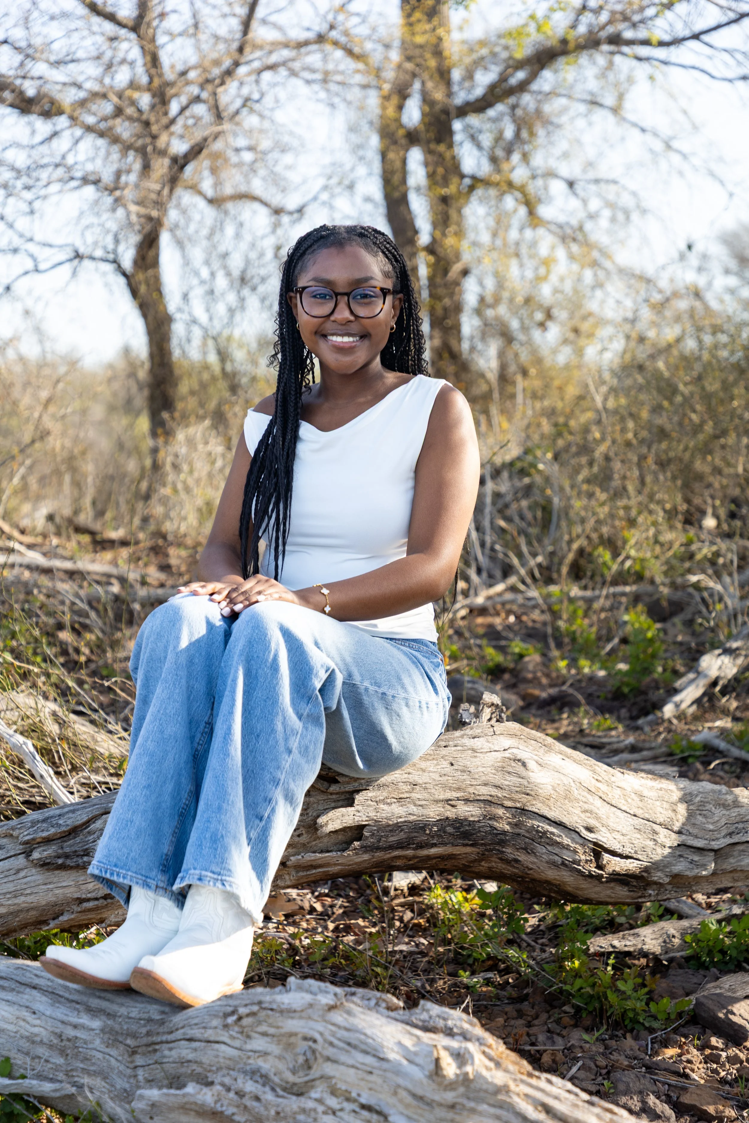 A smiling young woman with glasses, long braided hair, wearing a white sleeveless top, blue jeans, and white boots, sitting on a fallen branch in a natural outdoor setting with trees and light foliage.