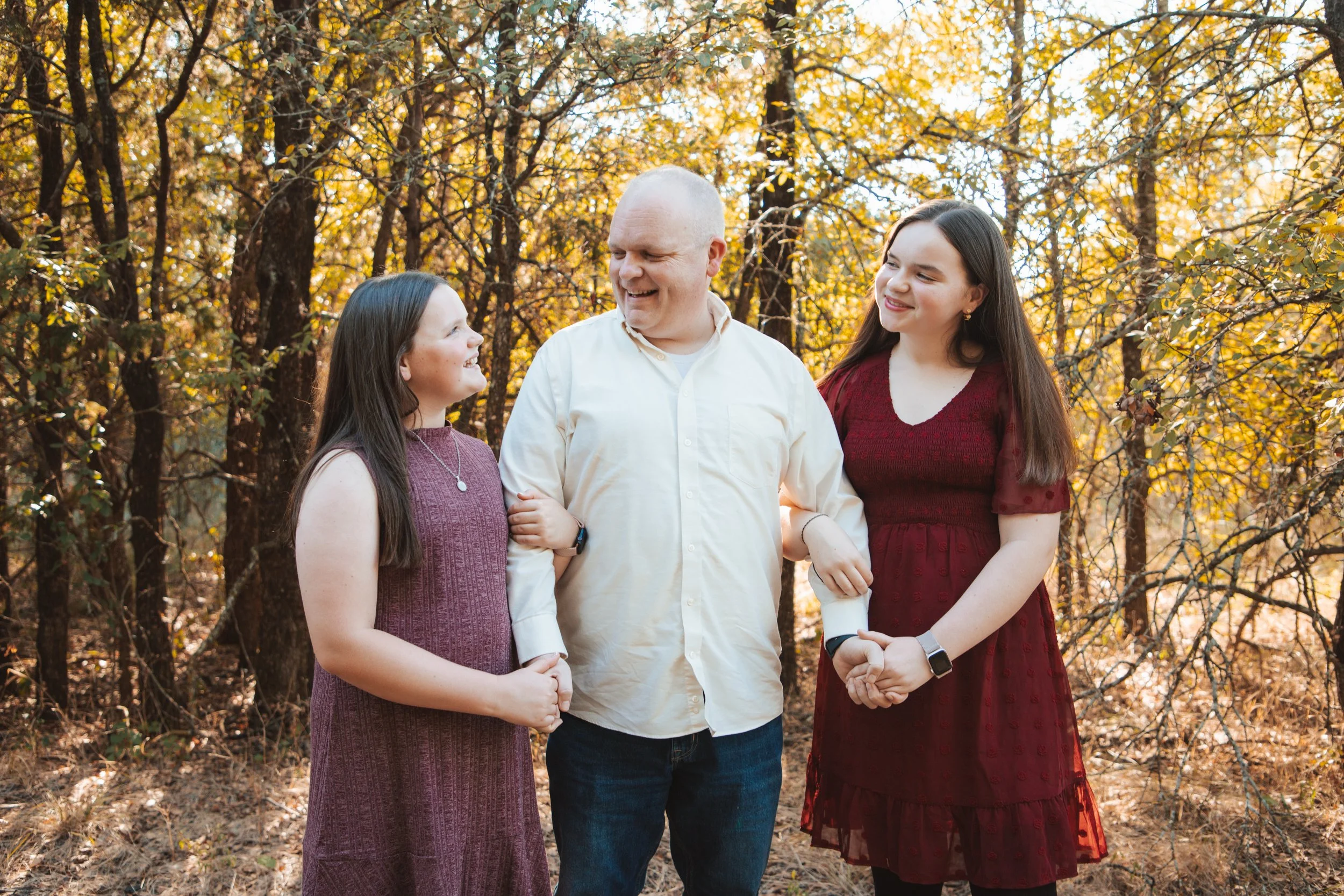 Family of four standing outdoors in a wooded area during fall, smiling and holding hands, with orange and yellow leaves.