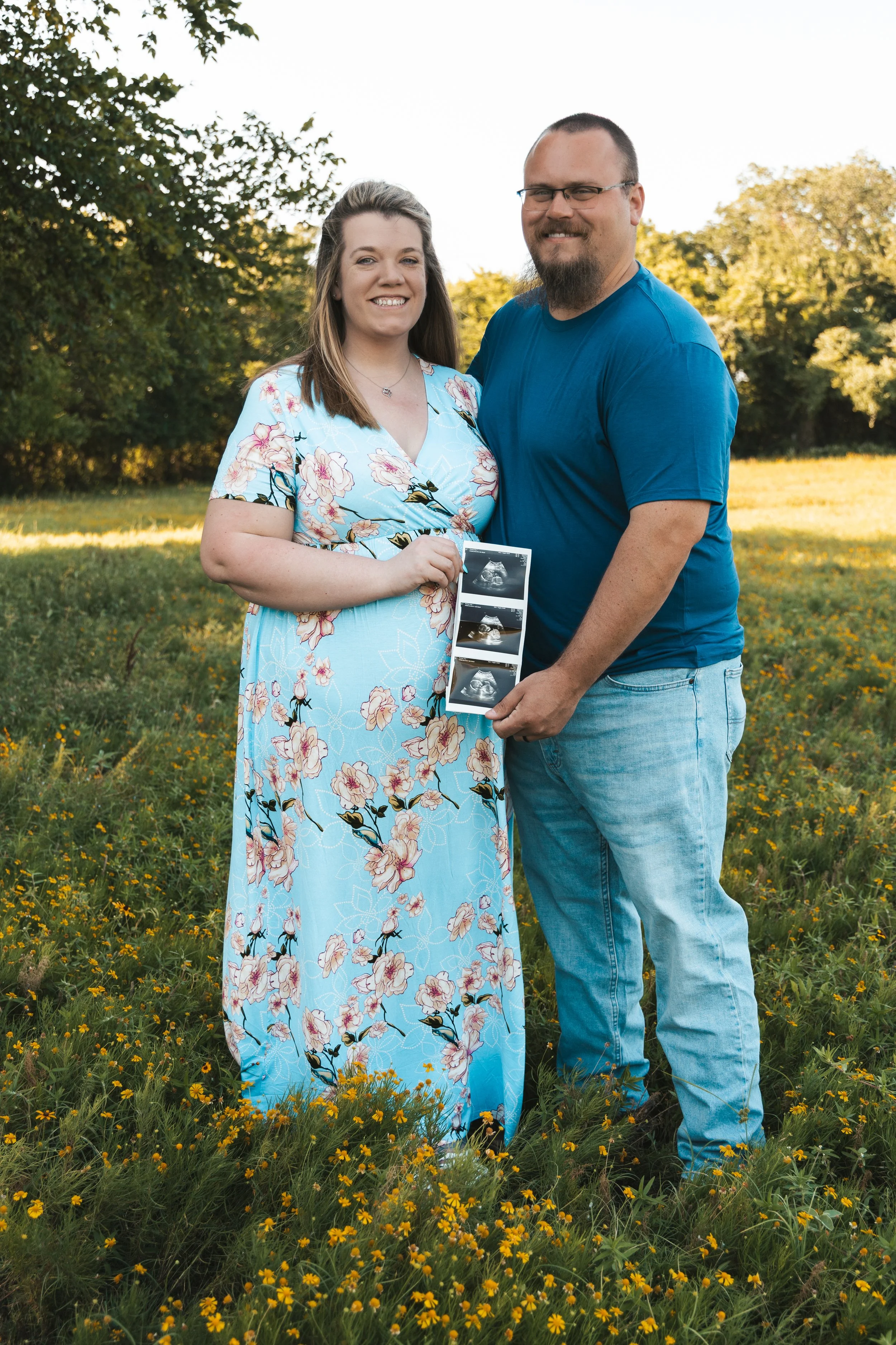 A couple stands outdoors in a field of yellow wildflowers, holding ultrasound images and smiling at the camera. The woman is wearing a light blue floral dress, and the man is dressed in a blue T-shirt and jeans. Trees are in the background.
