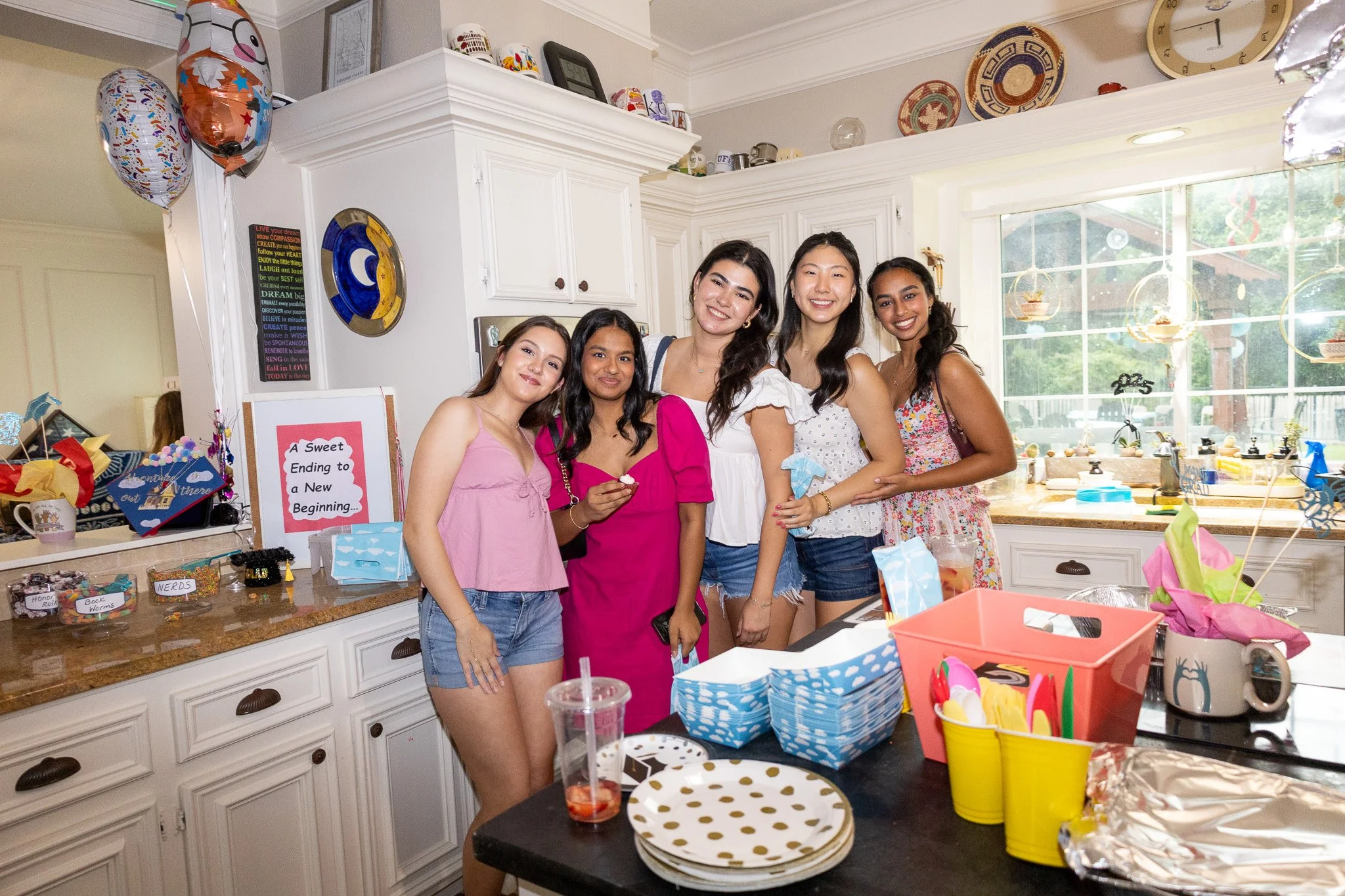 Group of five women standing together in a kitchen, celebrating a birthday. They are smiling, with colorful decorations, balloons, party favors, and a sign that reads 'A Sweet Ending to a New Beginning.'