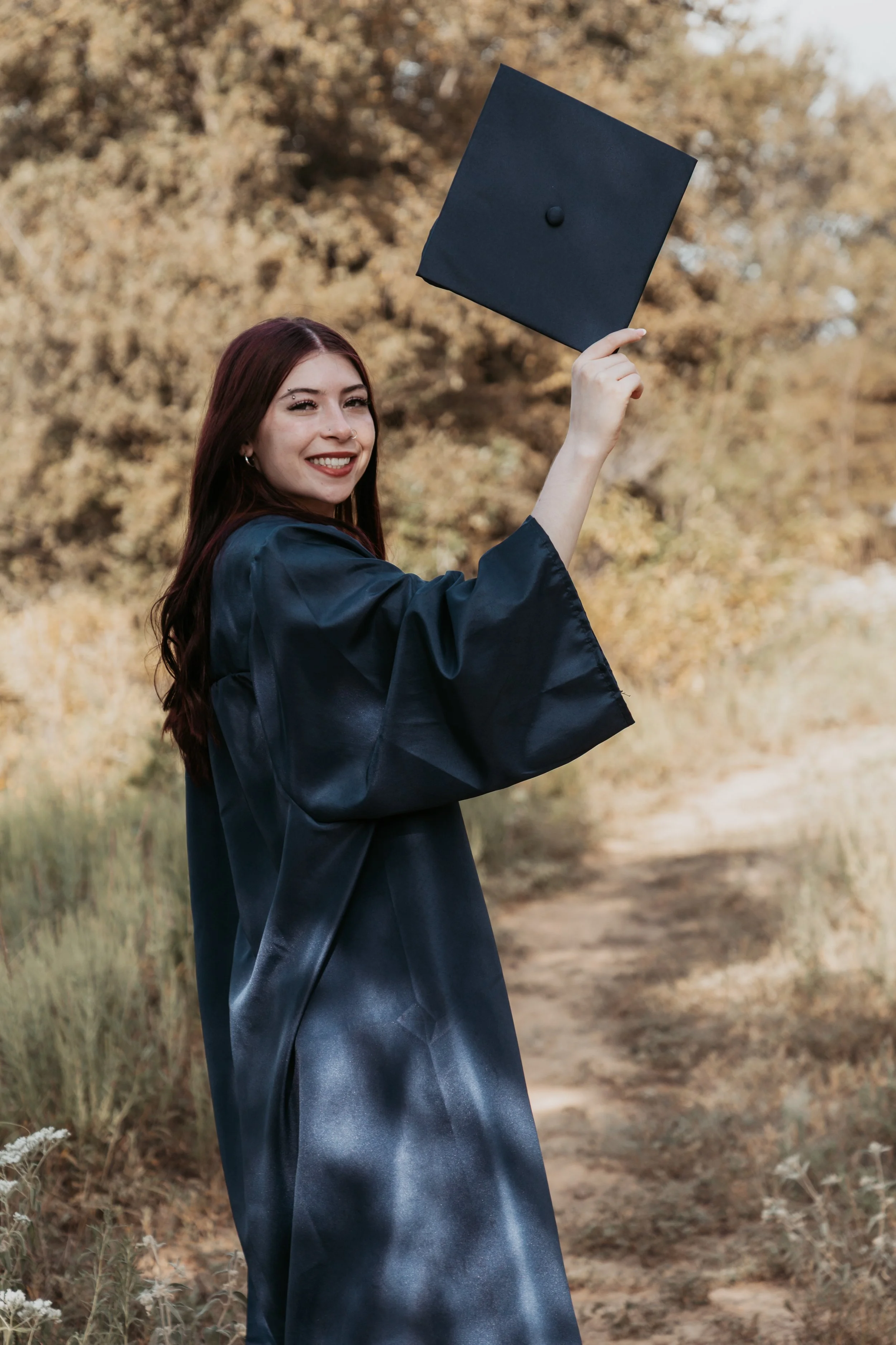 A young woman in a graduation gown holding a cap in an outdoor setting with trees in the background.