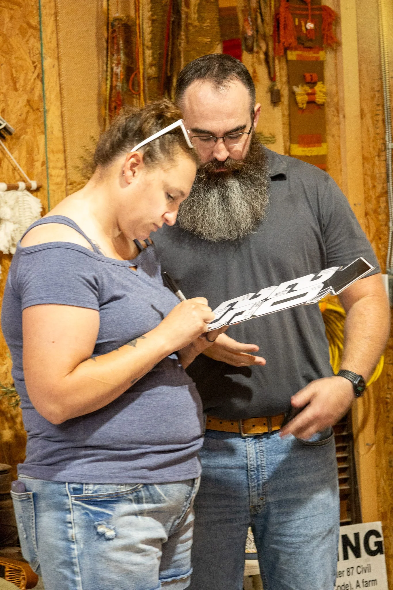 Two people standing in a workshop or rustic setting, looking at a sticker sheet together, one writing or pointing, the other watching.