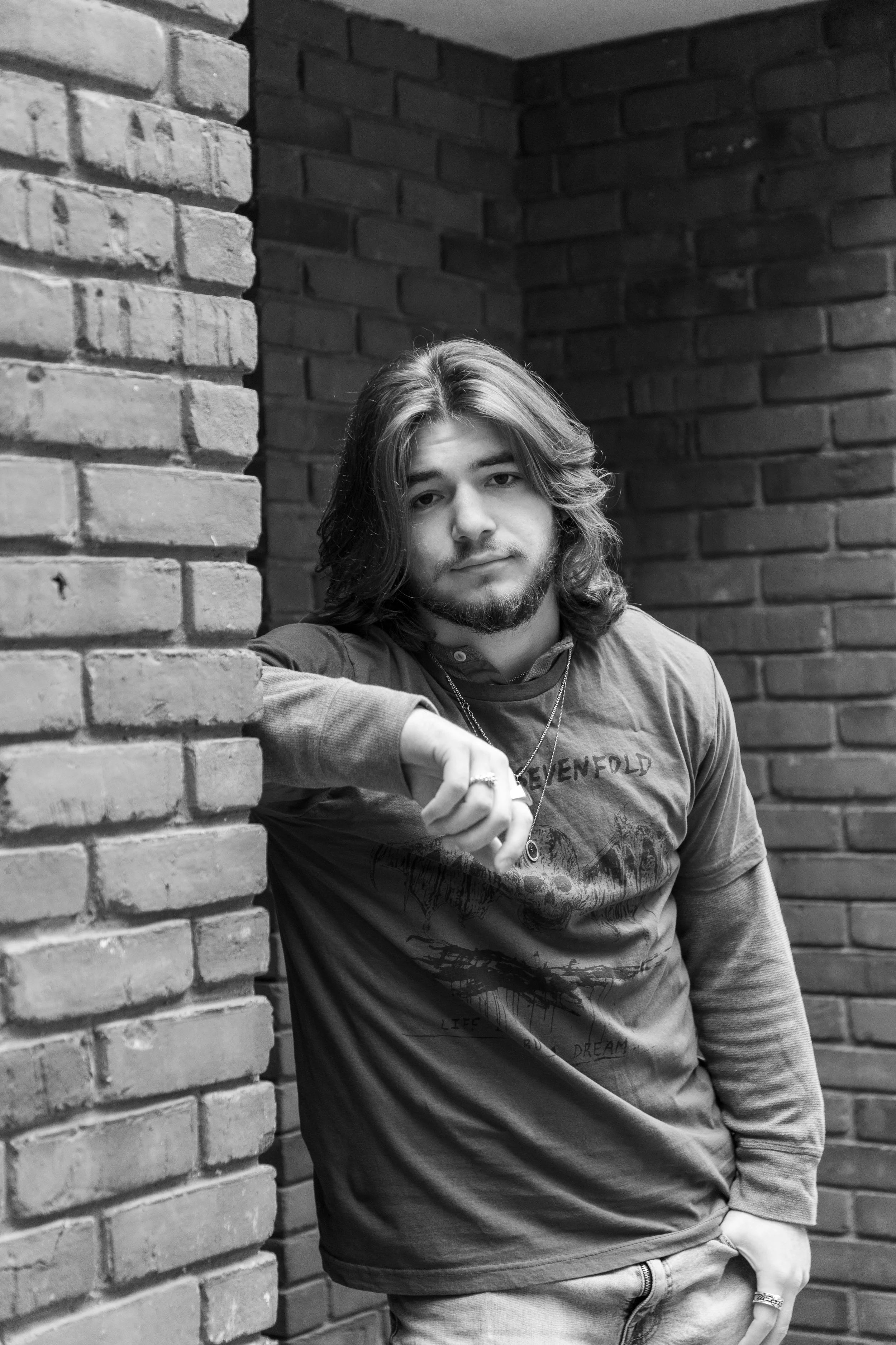 A young man with long hair, a beard, and a relaxed expression, standing against a brick wall and looking at the camera.