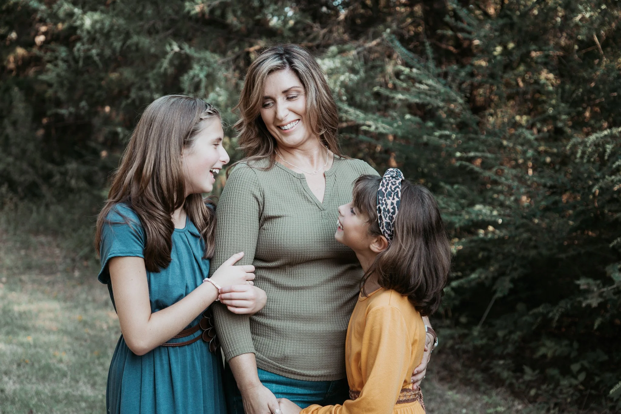 A woman with two young girls standing outdoors among trees, smiling and looking at each other.