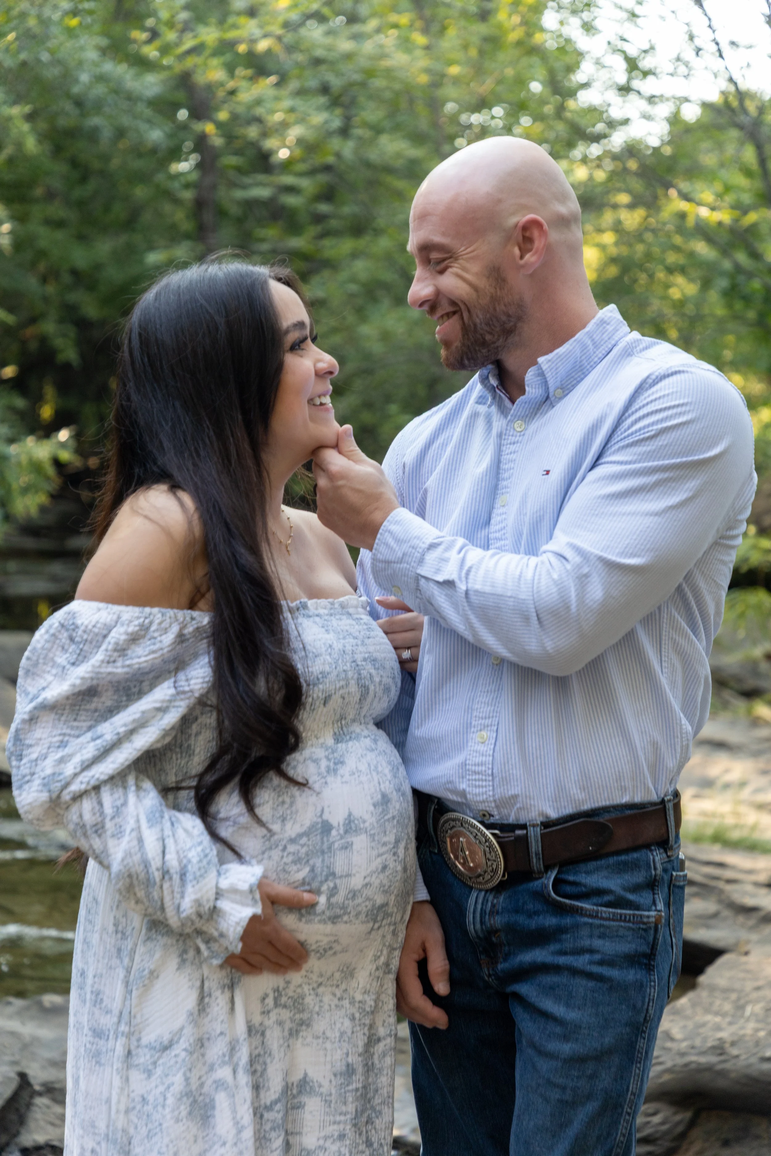 A pregnant woman and a man are smiling and looking at each other outdoors in a wooded area, with the man gently touching the woman's chin.