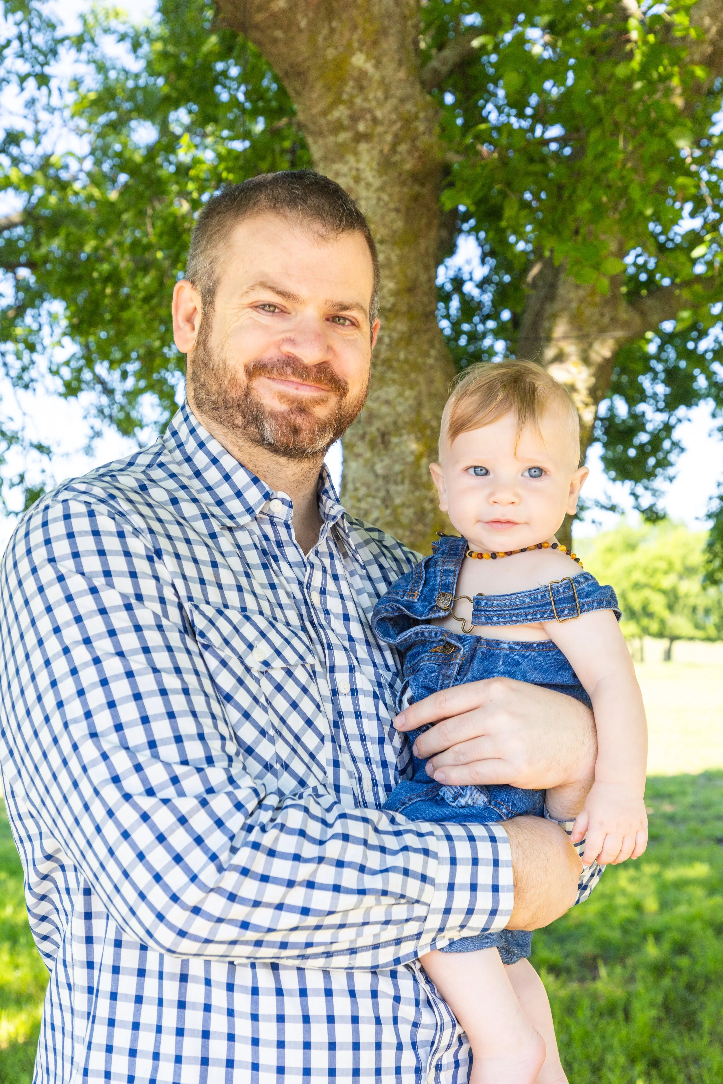 A man with a beard holding a young child outdoors near a large tree on a sunny day.