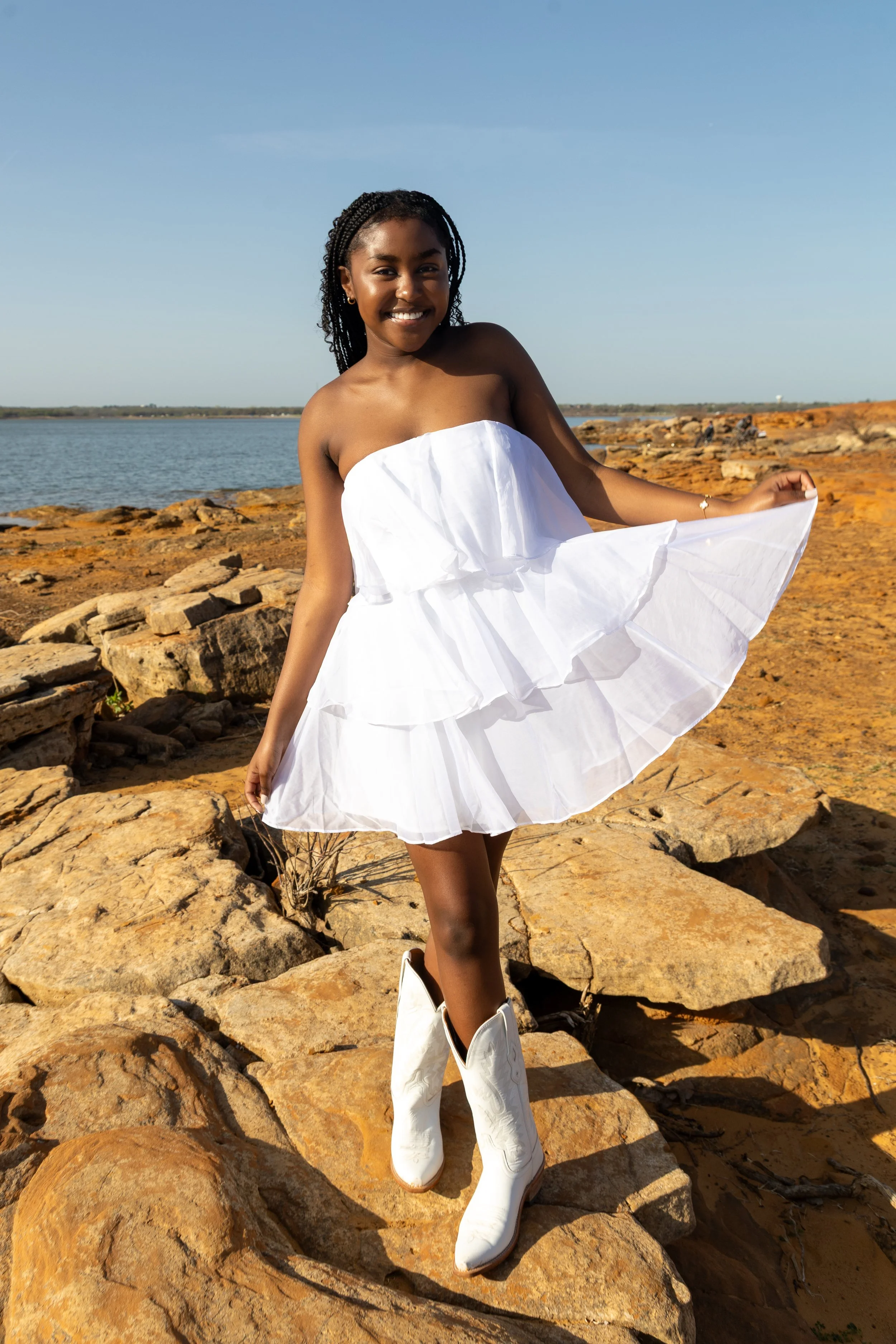 A smiling young girl in a white strapless dress and white cowboy boots standing on rocky terrain near water, with clear blue sky overhead.