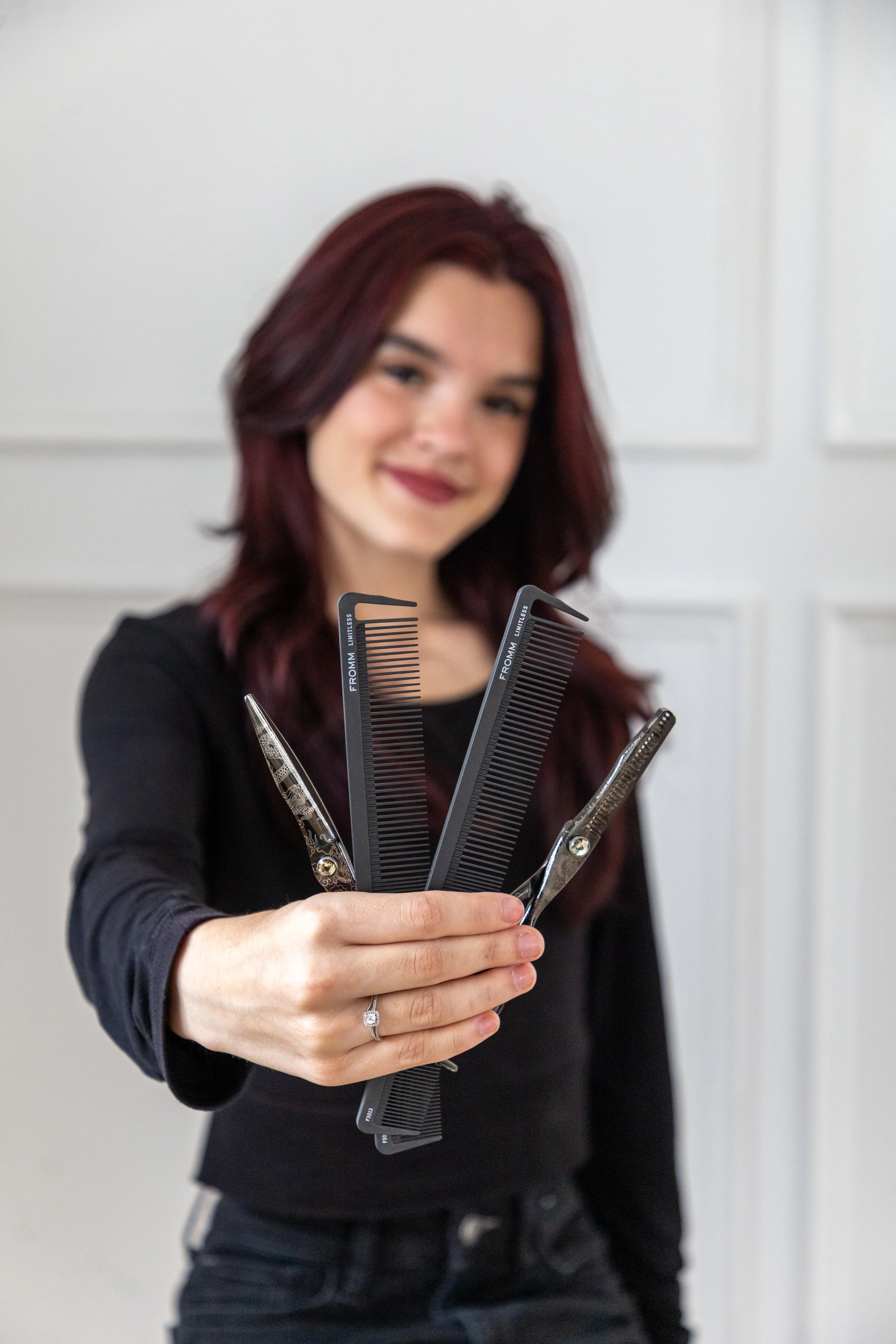 A woman with shoulder-length red hair wearing a black top, holding hair-cutting tools including scissors and combs, smiling and standing in front of a white background.
