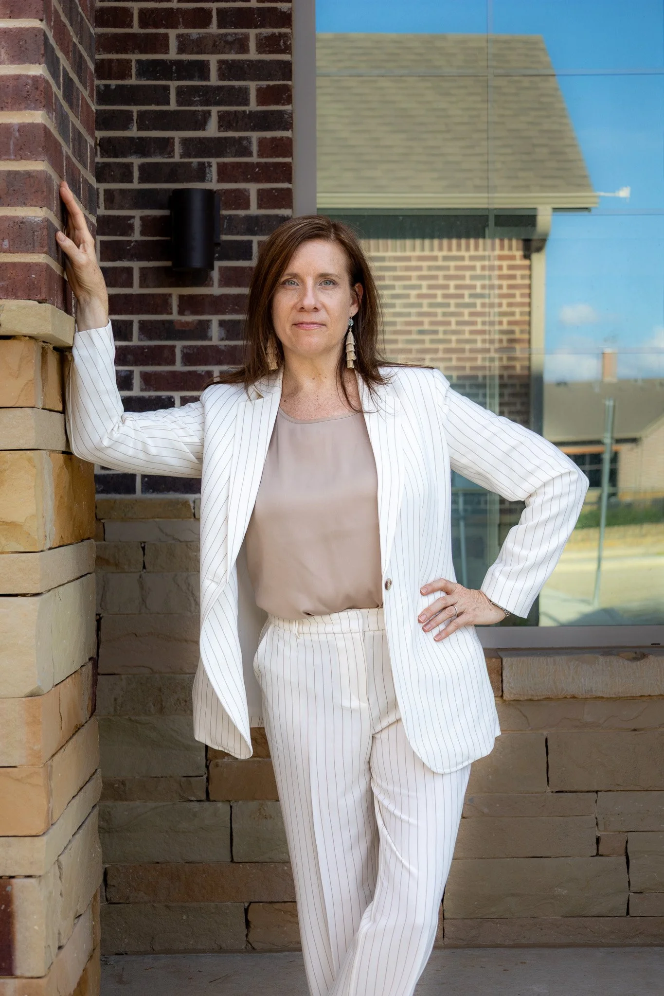 A woman in a white pinstripe suit standing outside near a brick wall and large window with a reflection of neighboring buildings.
