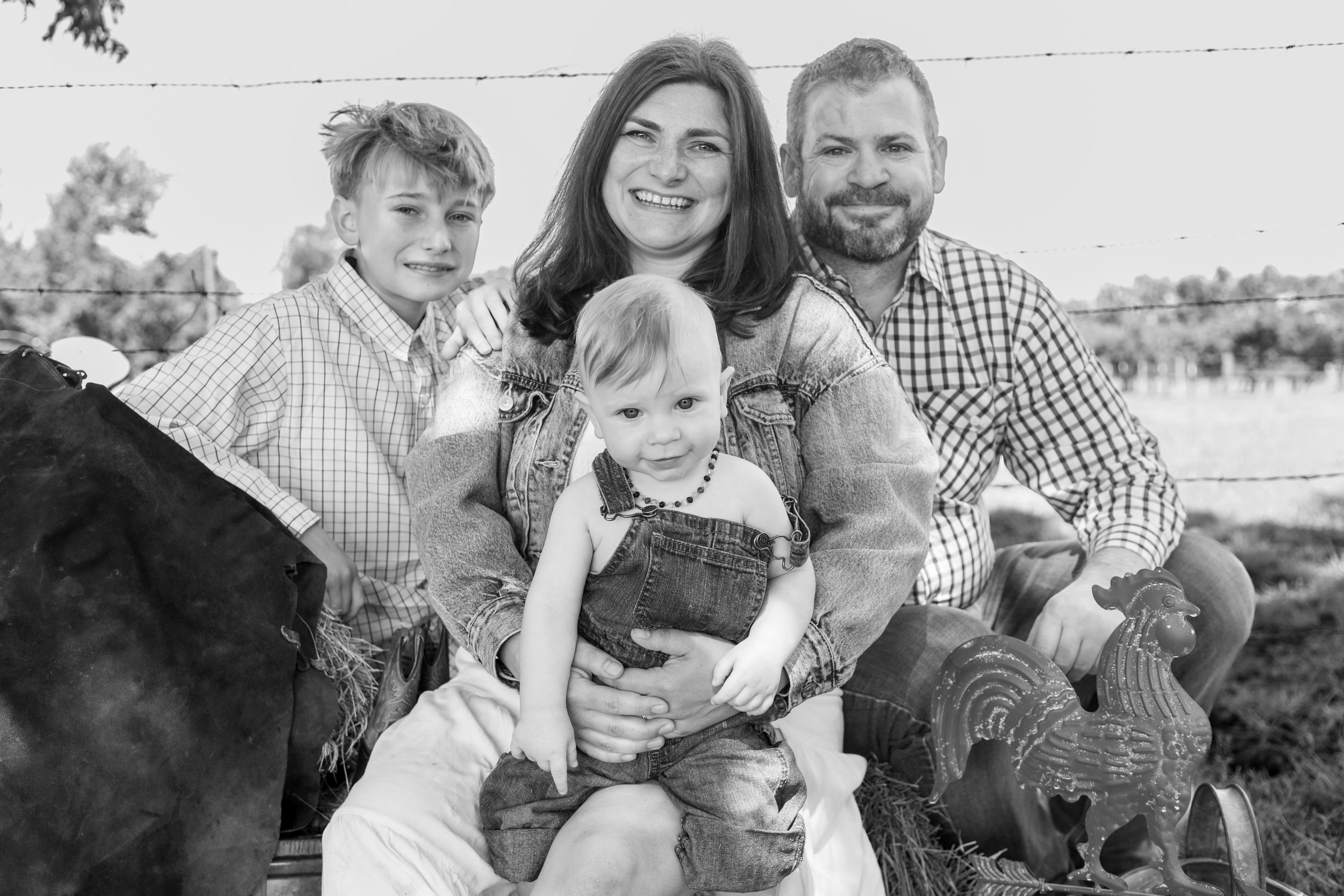 A family of five sitting outdoors next to a cow, with a rural background and barbed wire fence, smiling at the camera.