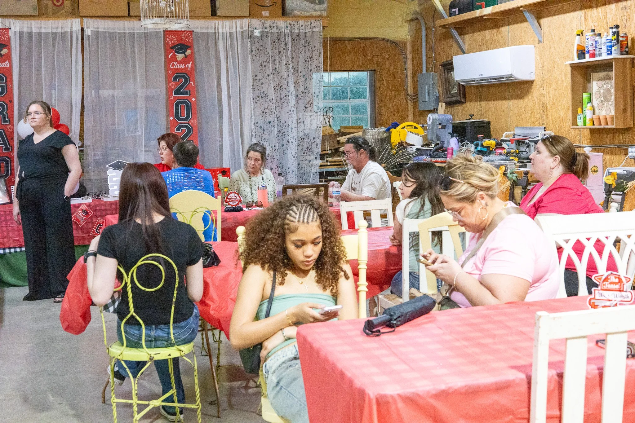 People attending a graduation celebration in a rustic indoor space with Halloween and graduation decorations, sitting at tables with red tablecloths.