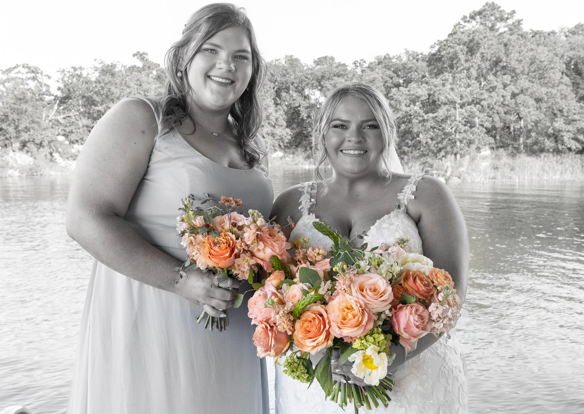 Two women, one in a white wedding dress and the other in a lighter dress, standing by a lake with trees in the background, holding colorful bouquets of flowers.