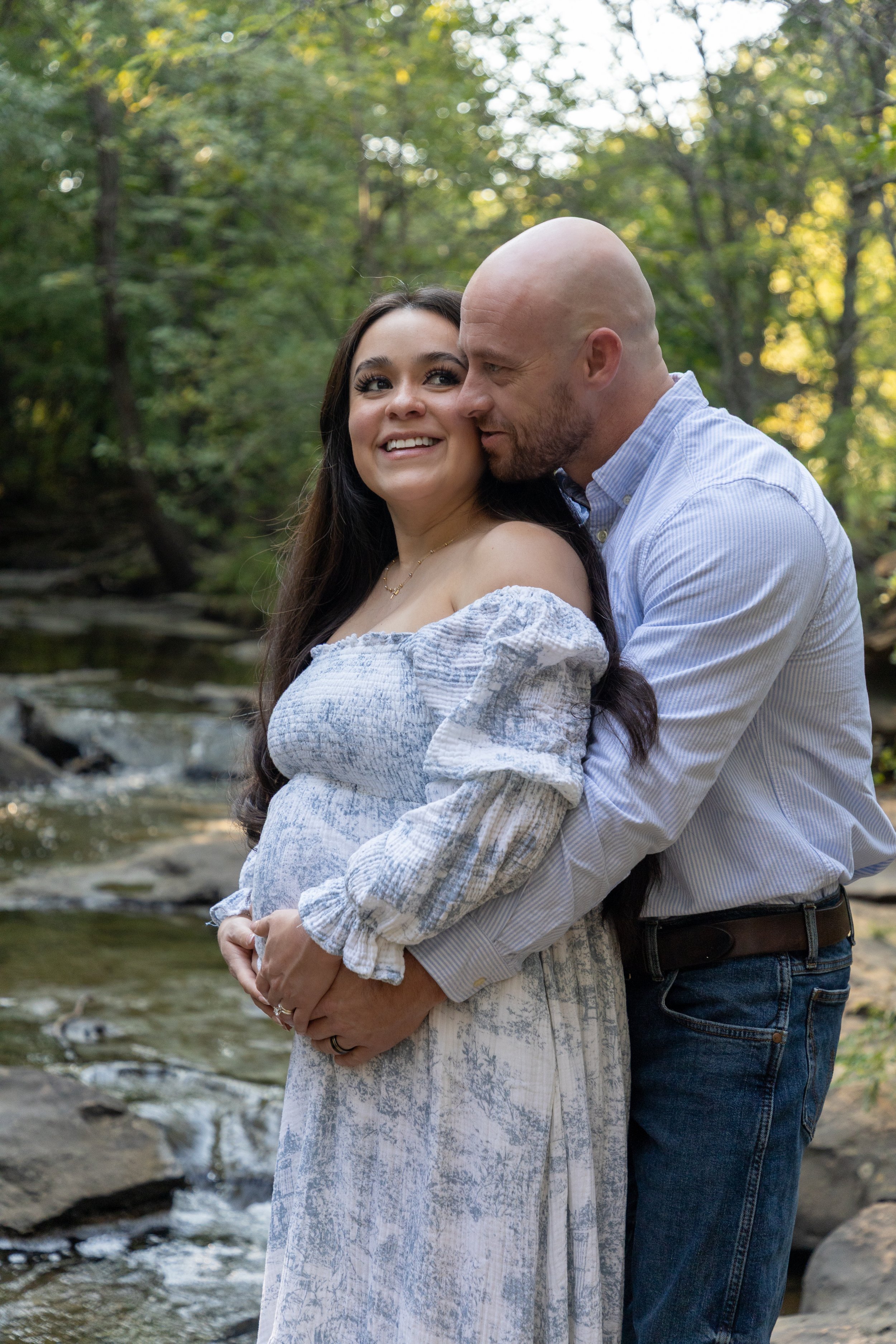 A couple standing by a stream in a wooded area, embracing each other. The woman is smiling and wearing an off-the-shoulder dress, while the man is kissing her cheek.
