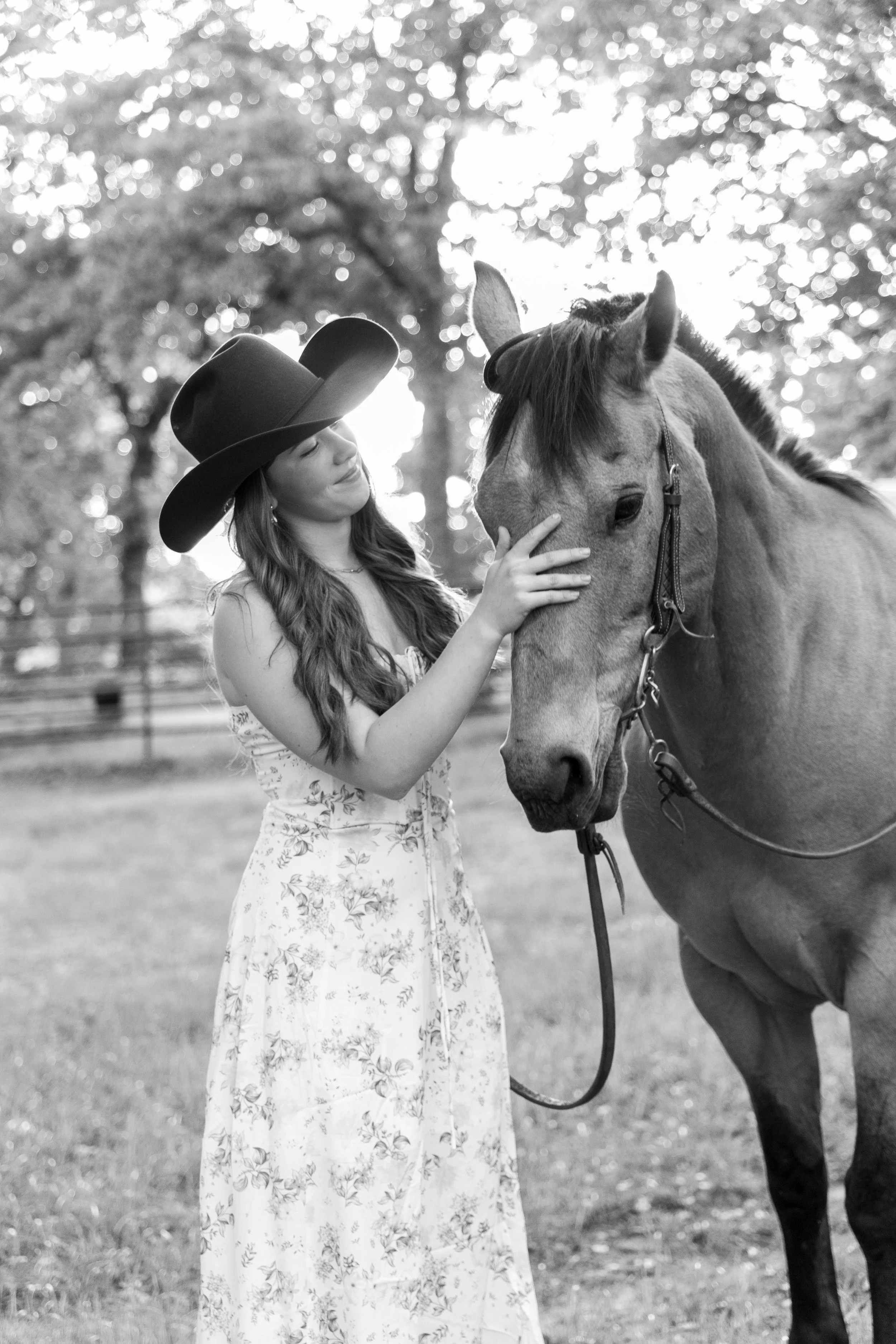 A girl wearing a wide-brimmed hat and a floral dress gently touching a horse outdoors with trees and sunlight in the background.