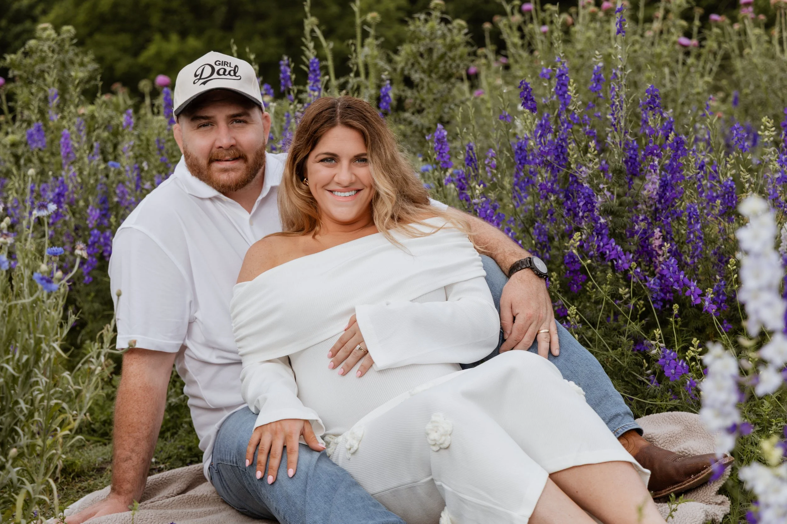 A smiling couple sitting on blankets in a field of purple flowers, with the woman leaning back on the man's lap, both looking at the camera.