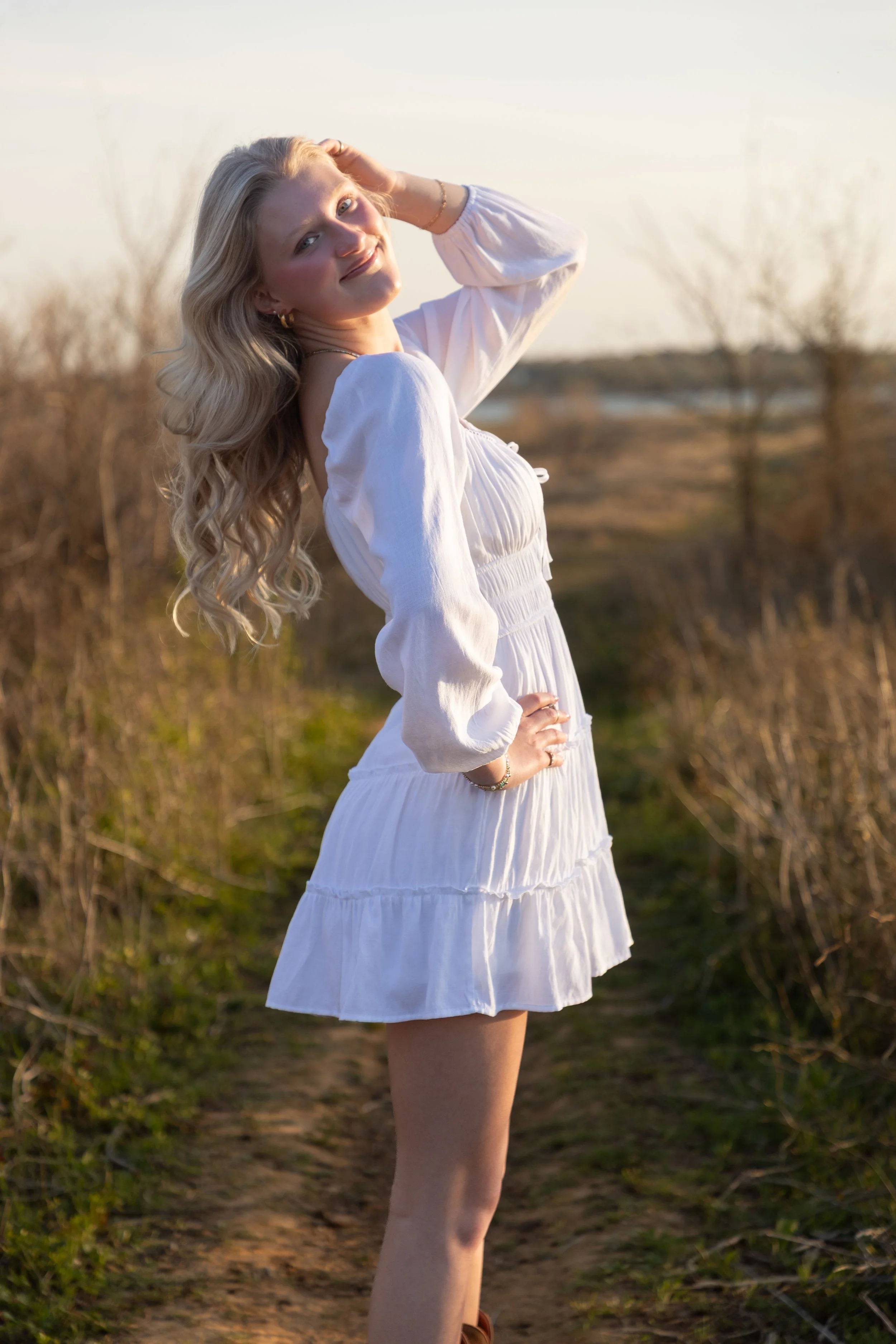 A young woman with long blonde hair, dressed in a white dress, posing on a dirt path outdoors during sunset or late afternoon.