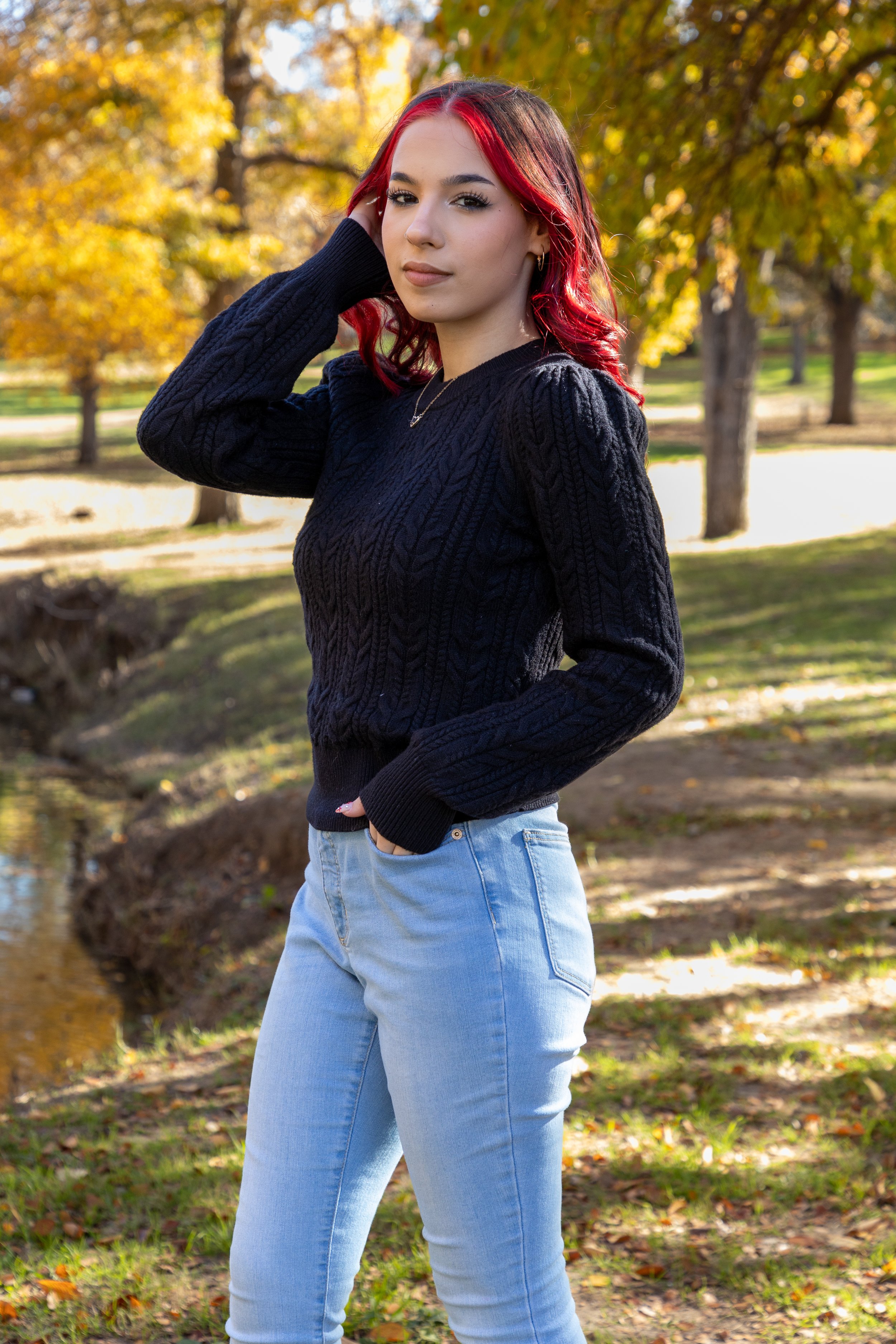 A young woman with red hair, wearing a black sweater and light blue jeans, standing outdoors in a park with autumn trees in the background.