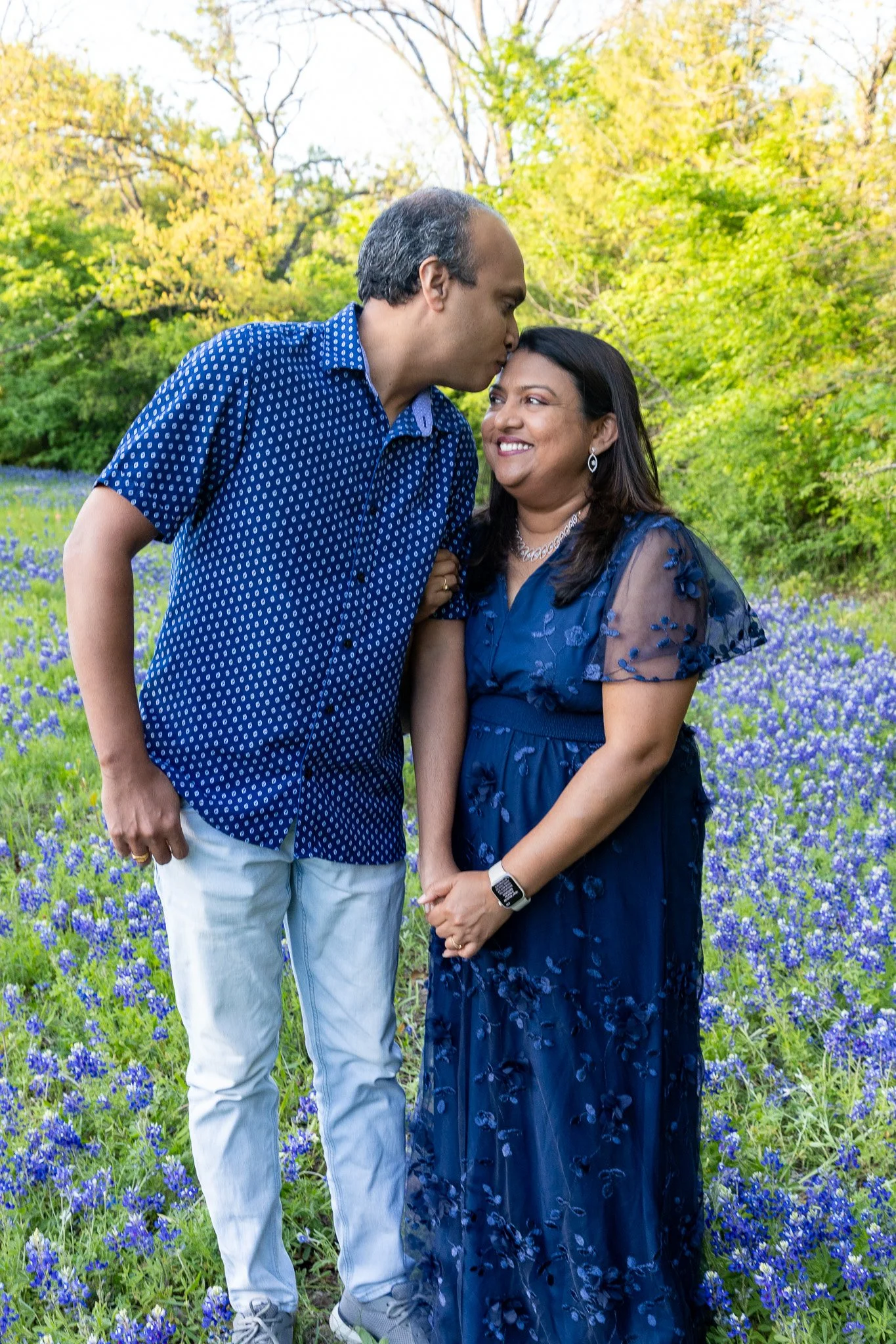 A man with dark hair, wearing a blue patterned shirt and light-colored pants, kisses a woman on the forehead. The woman has dark hair, wearing a dark blue dress with floral details, jewelry, and a smartwatch. They are standing outdoors in a field of 