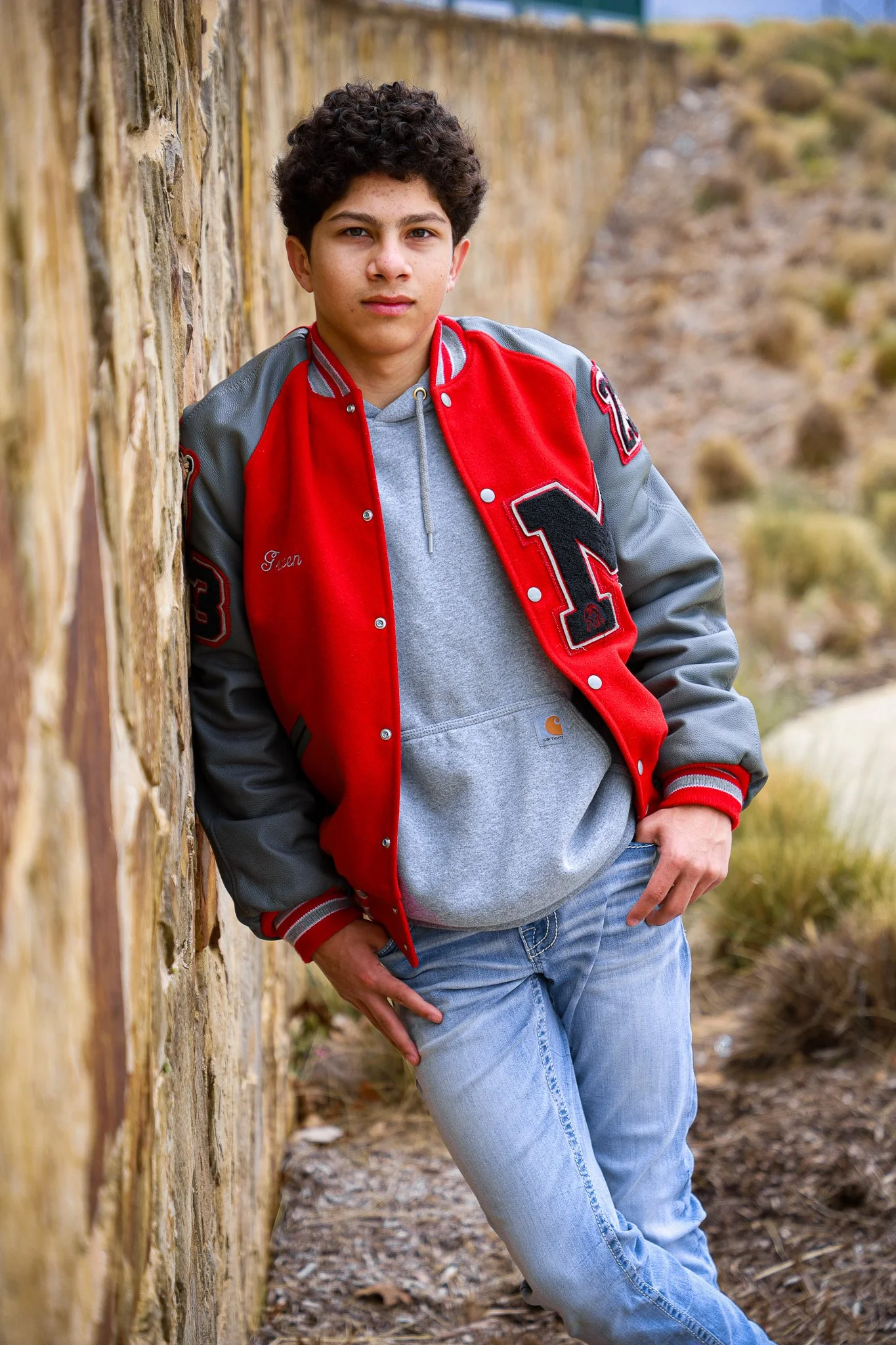 A teenage boy with curly dark hair leaning against a stone wall outdoors. He is wearing a red and gray varsity jacket, a gray hoodie, and light blue jeans, with a desert landscape in the background.