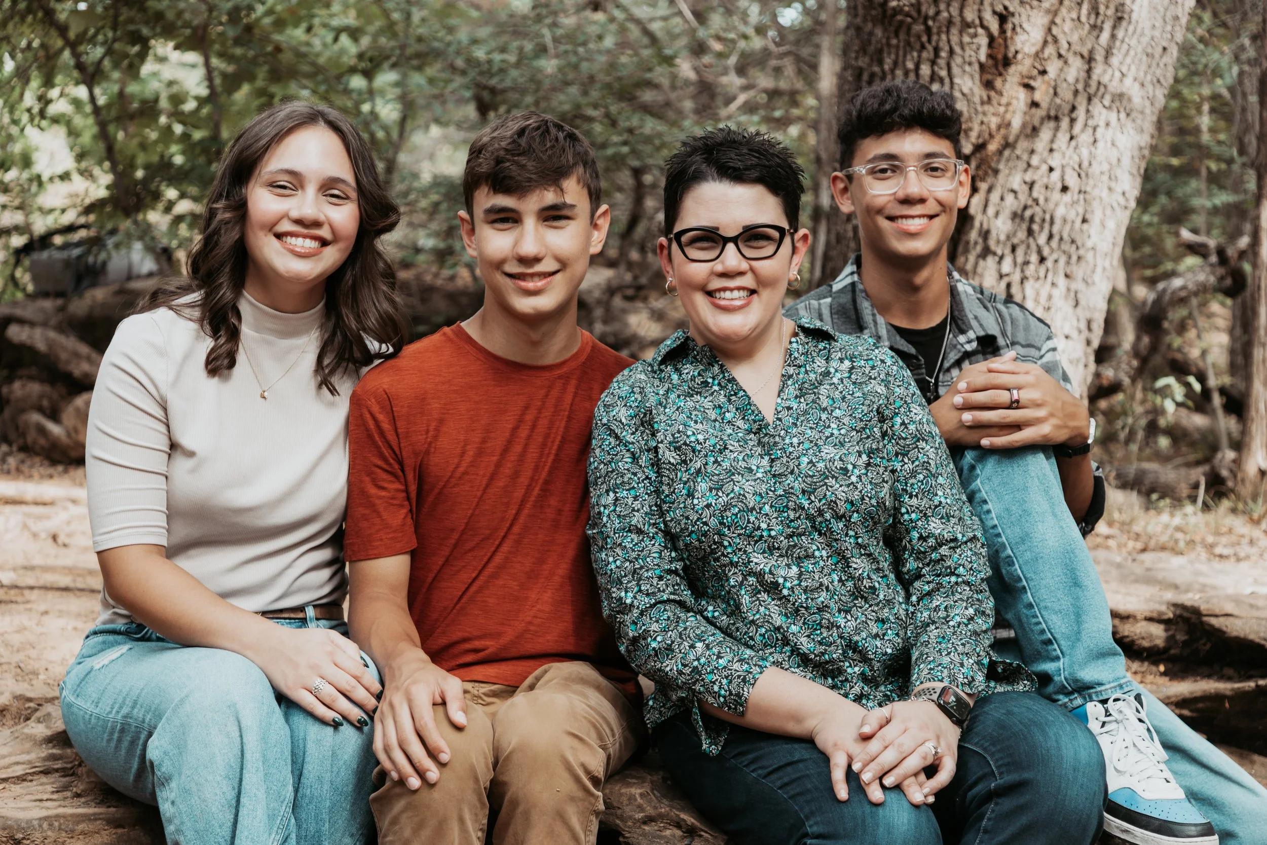 A group of five smiling people sitting on a log outdoors in a forest.