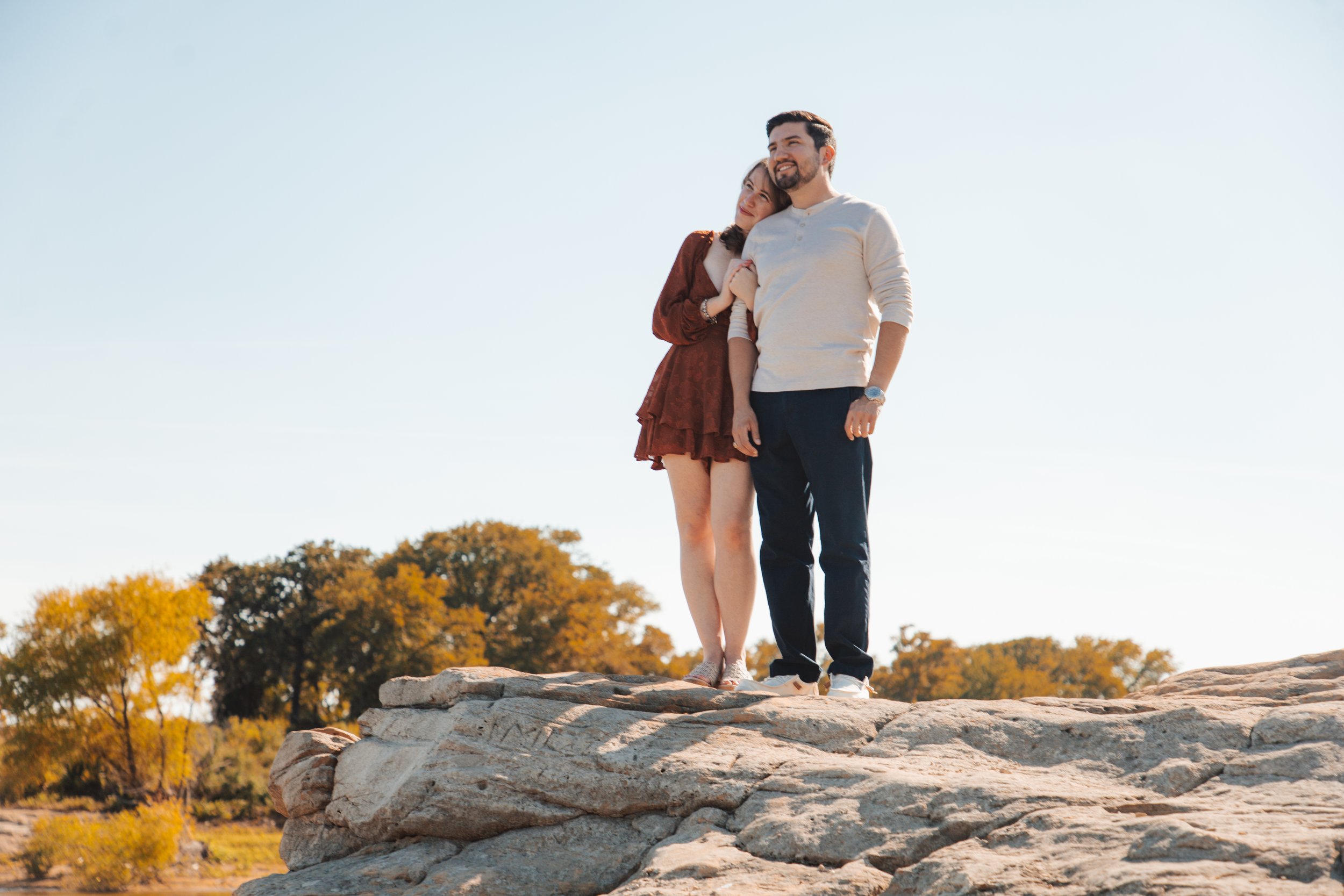 A couple stands on a rocky ledge outdoors, with the woman resting her head on the man's shoulder and holding his arm. The background features trees with autumn foliage and a clear sky.
