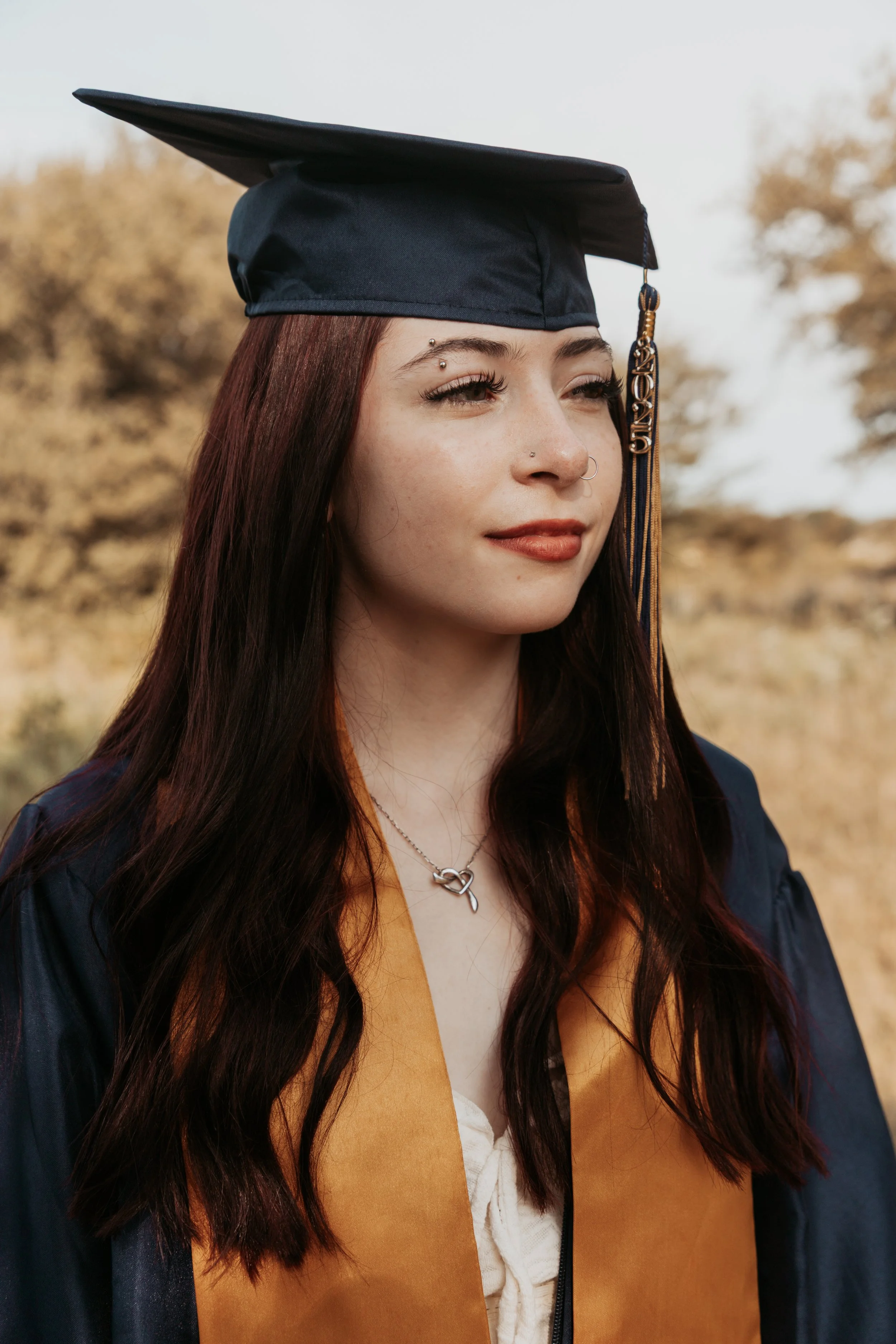 A young woman in a navy blue graduation cap and gown, with long dark hair, standing outdoors with trees in the background.