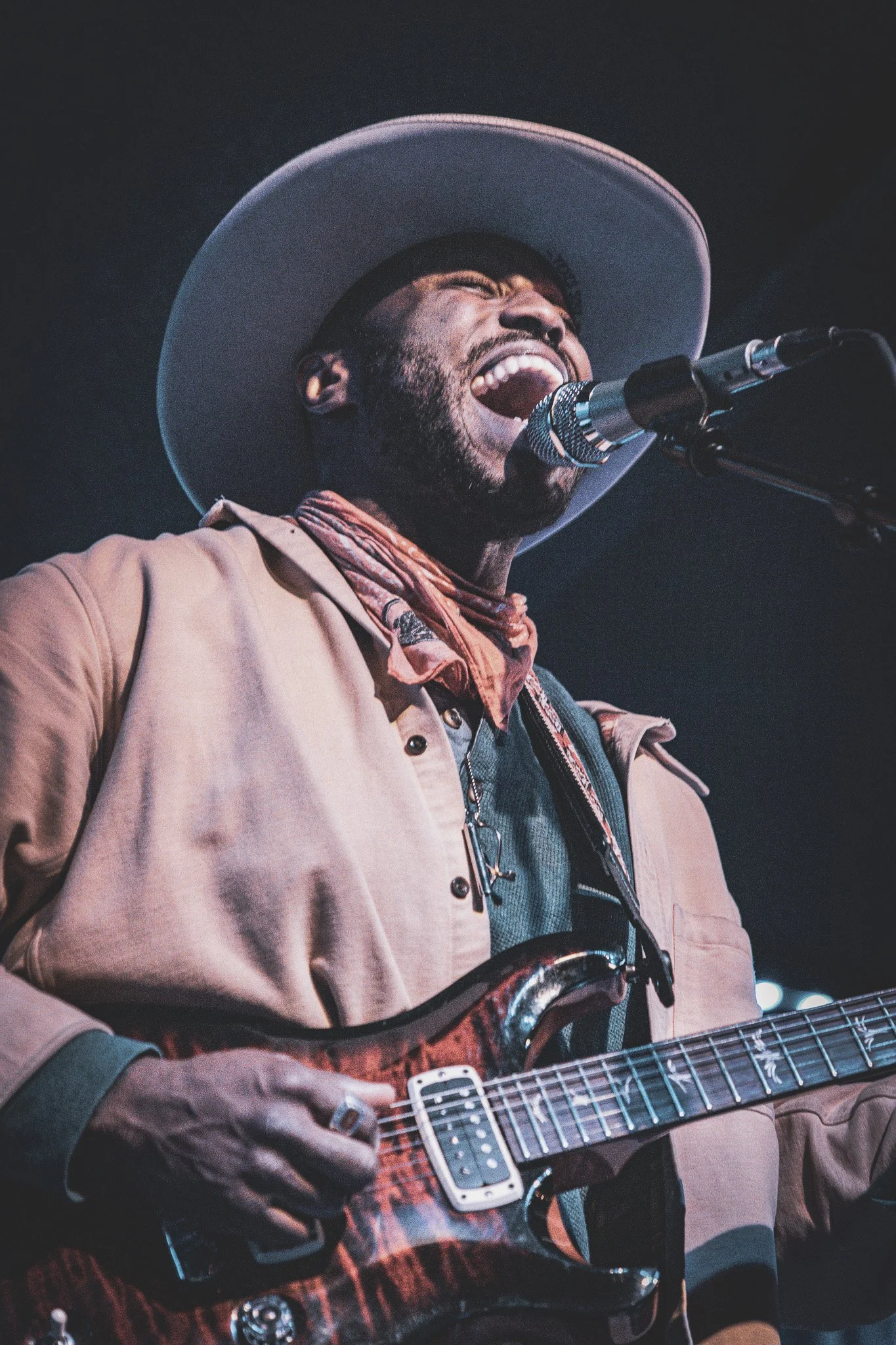A man wearing a large hat plays an electric guitar and sings into a microphone on stage.
