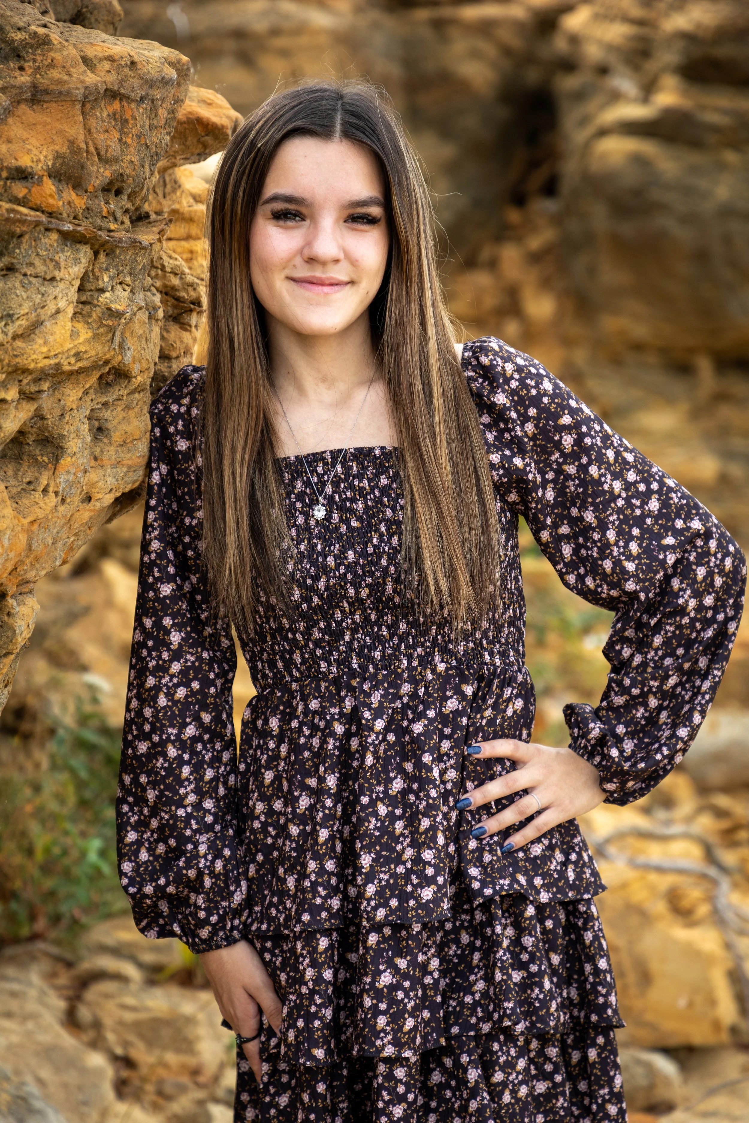 A young woman with long brown hair wearing a black floral dress standing outdoors near rocks.
