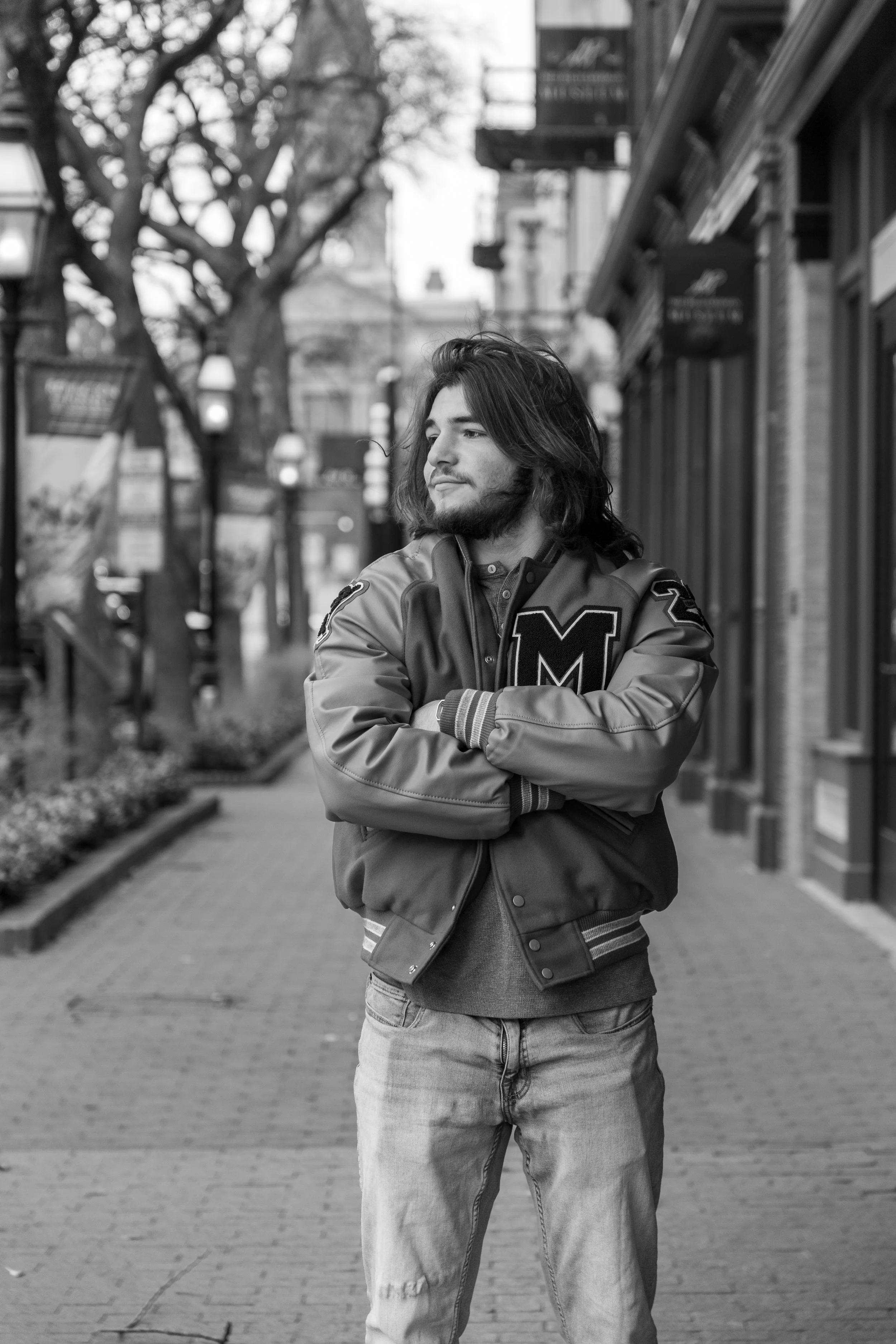 Black and white photo of a young man with long hair and a beard standing on a city sidewalk, crossing his arms and looking off to the side. He is wearing a varsity jacket with an 'M' patch.